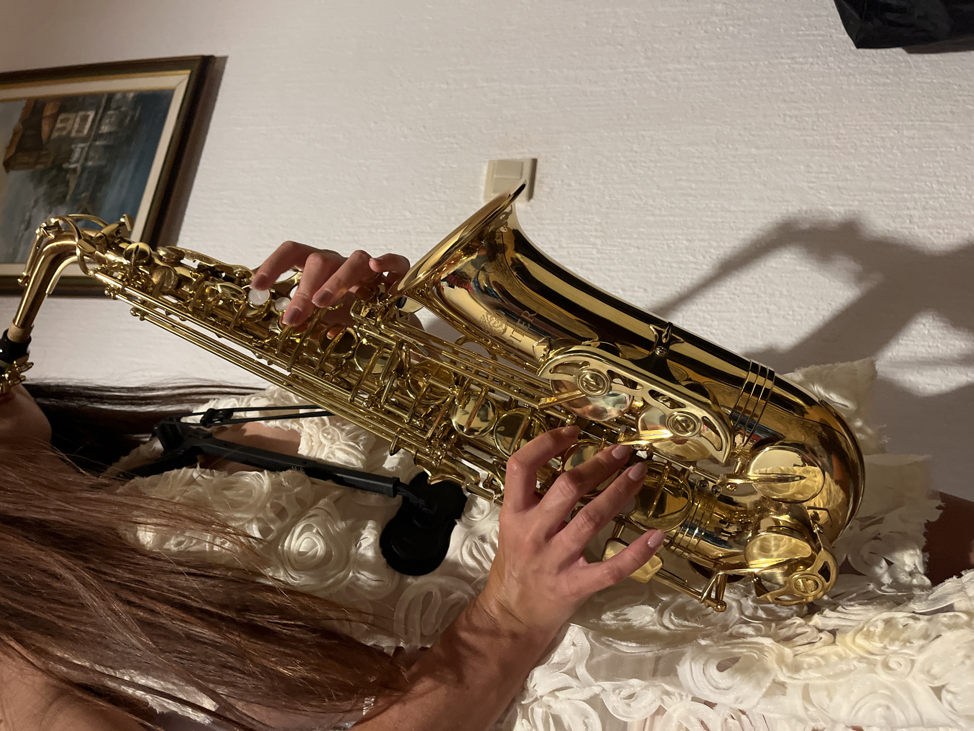 A woman with long, dark brown hair is playing a golden saxophone. She is wearing a white dress with a textured, floral-like pattern. Her hands are positioned on the keys of the saxophone. The background shows a textured white wall with a framed painting hanging on it and a light switch. The scene is indoors, with some shadows cast on the wall. The location is identified as Šušanj, Montenegro.