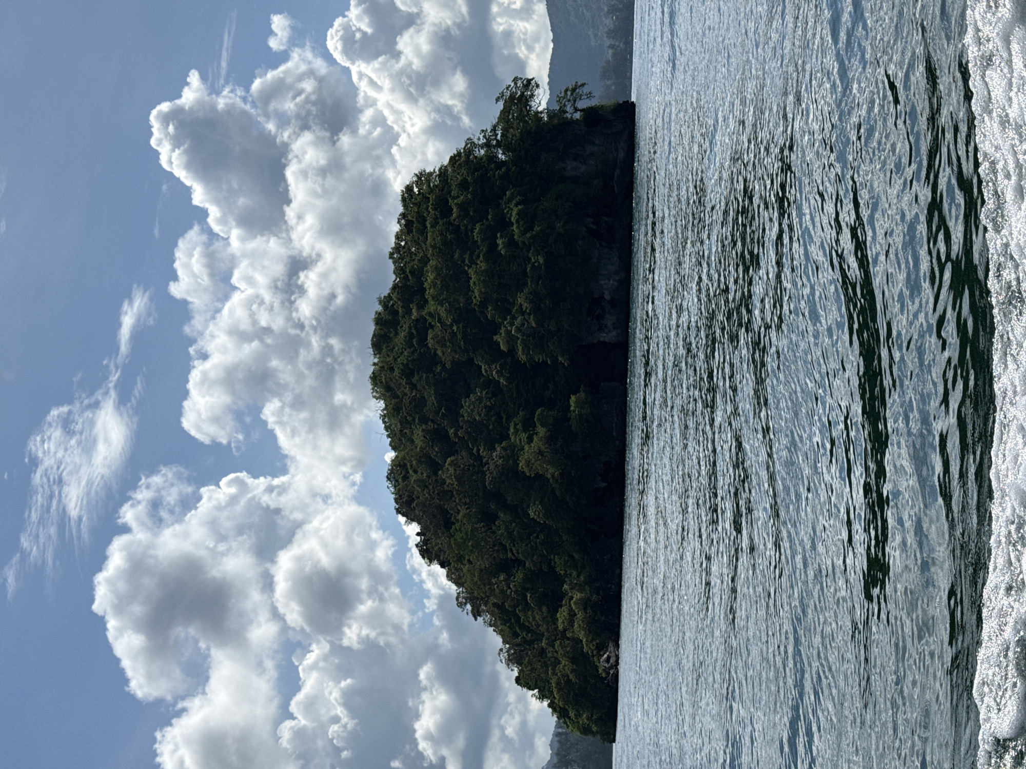The image shows a lush green island with trees on a body of water. The sky above is blue with scattered white clouds. The water is rippling and reflects the light. In the foreground, there is a wake of white foam, indicating movement in the water. The island appears to be uninhabited. There are no people visible in the image. The scene is natural and scenic, likely a tropical or subtropical location. There is no indication that this location is in Ban Klong Haeng, Thailand.