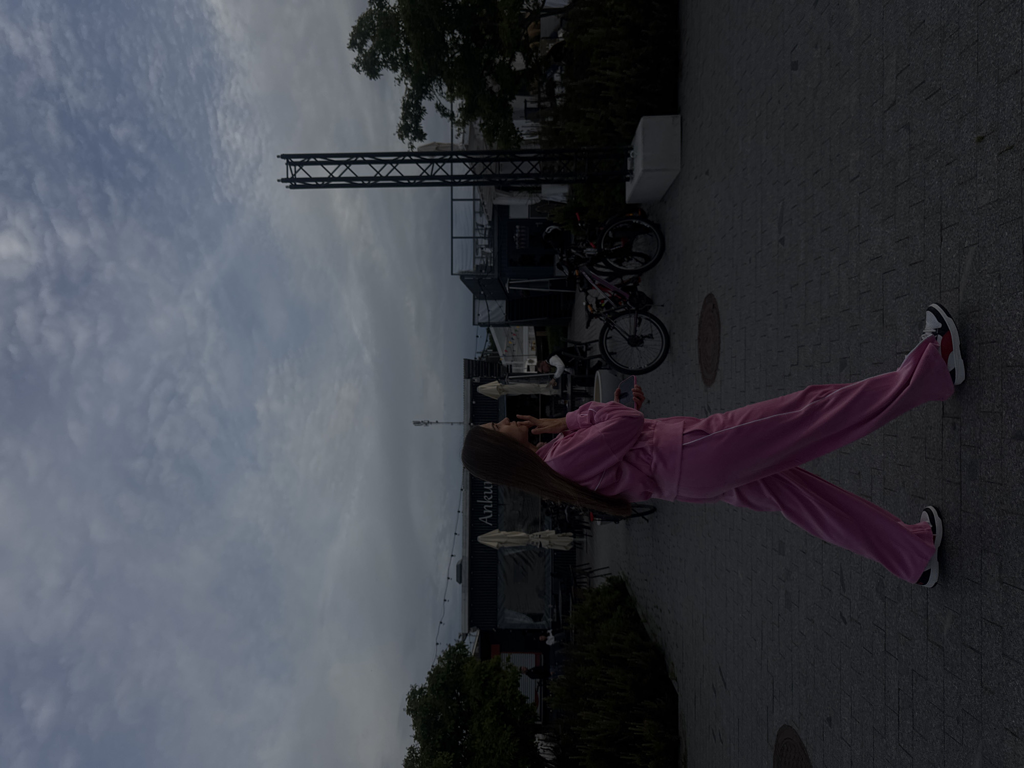 A woman in a pink tracksuit walks on a paved pathway. She is looking to her right and holding her phone to her ear. In the background, there are two bicycles parked near a building with outdoor seating and umbrellas. There is also a tall metal structure on the right side of the image and trees on the left. The sky is cloudy. The scene appears to be an outdoor dining or recreational area in Tallinn, Estonia.