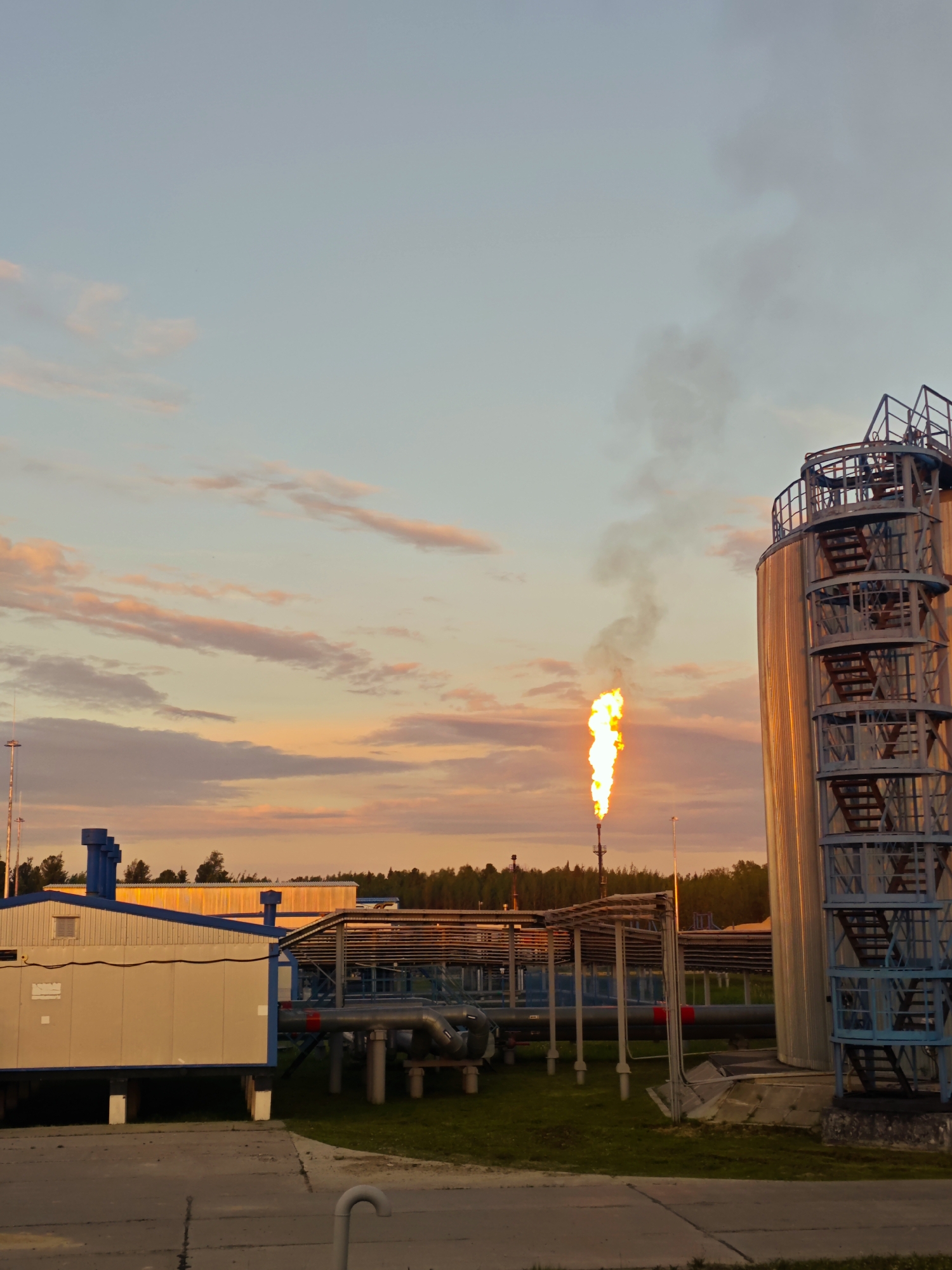 This is a vertical, eye-level, full shot of an industrial site at dusk. The sky is a pale blue with soft pink and orange clouds towards the horizon. A large, cylindrical metal tank with external stairs is on the right side of the frame, reflecting the sunset. To the left of the tank, a tall flare stack emits a bright orange flame and a plume of gray smoke. In the mid-ground, a long, low building with a white and tan facade and blue trim sits on stilts. Behind it, numerous pipes and structures stretch across the landscape, supported by metal frameworks. A dense line of trees forms the horizon in the background, silhouetted against the colorful sky. The ground in the foreground is paved concrete. There are no people visible in the image.