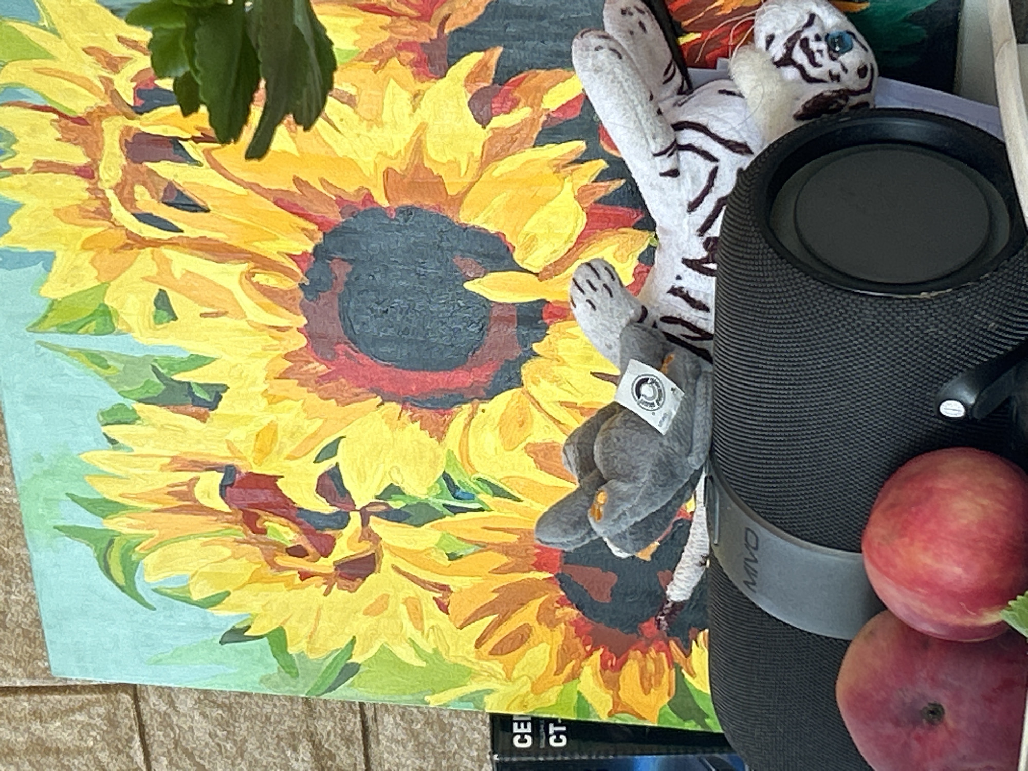 The image shows a close-up of a painting of sunflowers, with a black portable speaker in the foreground. Two stuffed animals, a gray cat and a white tiger, are resting on top of the speaker. Two peaches are also visible in the lower left corner of the image. The painting is on a light blue background with green leaves and yellow sunflowers. The speaker has the brand name "MIVO" visible. The scene appears to be indoors, possibly on a shelf or table, with a brick wall visible in the background. There are no people or actions depicted in the image. The location within the city of Калицино, Russia is not discernible from the image itself.
