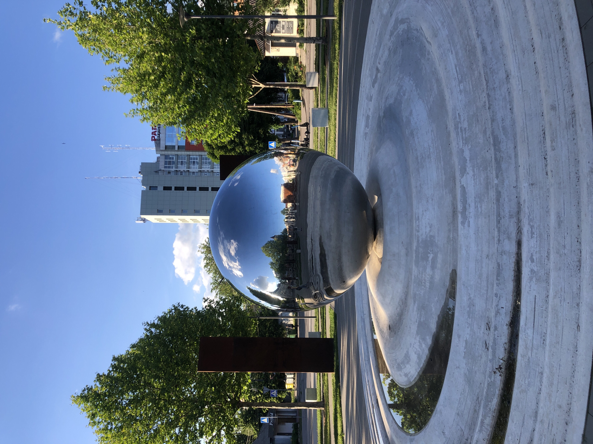 The image presents a bright daytime scene centered around a large, highly reflective, spherical metallic sculpture or fountain. This polished sphere acts as a convex mirror, reflecting an inverted and distorted panoramic view of its surroundings. It rests within a light gray, textured concrete basin or structure that exhibits concentric circular patterns and appears weathered or stained with darker patches, possibly from water.
The reflections in the sphere reveal a street scene, including buildings, numerous green trees, and what seem to be parked scooters or bicycles along a sidewalk. A blue triangular road sign, likely a yield sign, is clearly visible in the reflection, as is faint text that appears to read "Pihlaka" on a building or sign.
Beyond the sculpture in the actual scene, there are several multi-story buildings under a clear, bright blue sky with scattered white clouds. One prominent building features a red and white sign displaying the text "PAI OFFICE". Lush green deciduous trees line the street, suggesting a spring or summer season. Streetlights, power lines, and antennae are visible against the sky, along with a dark, rectangular sign or barrier near the trees.
The scene depicts a public urban or suburban space. The clear blue sky and strong sunlight indicate a sunny day, likely morning or afternoon. No people are performing any specific activity, contributing to a static and serene atmosphere. The image is oriented incorrectly, rotated 90 degrees counter-clockwise from its likely intended upright view.