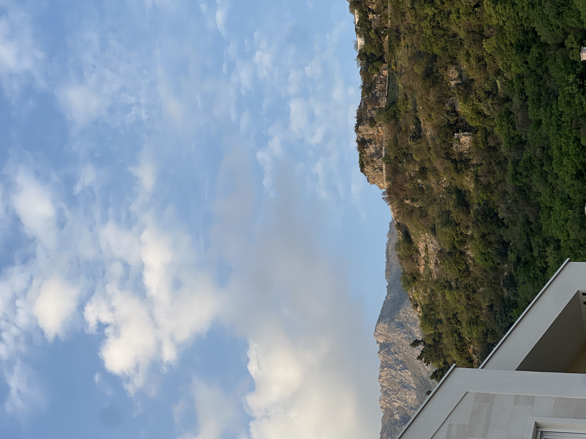 The image shows a view in Budva, Montenegro, with a bright blue sky and scattered white clouds above a mountain range covered in green trees. The perspective is upward, from a low angle, showing the corner of a modern white building in the lower right. The mountain has rocky cliffs and dense vegetation.