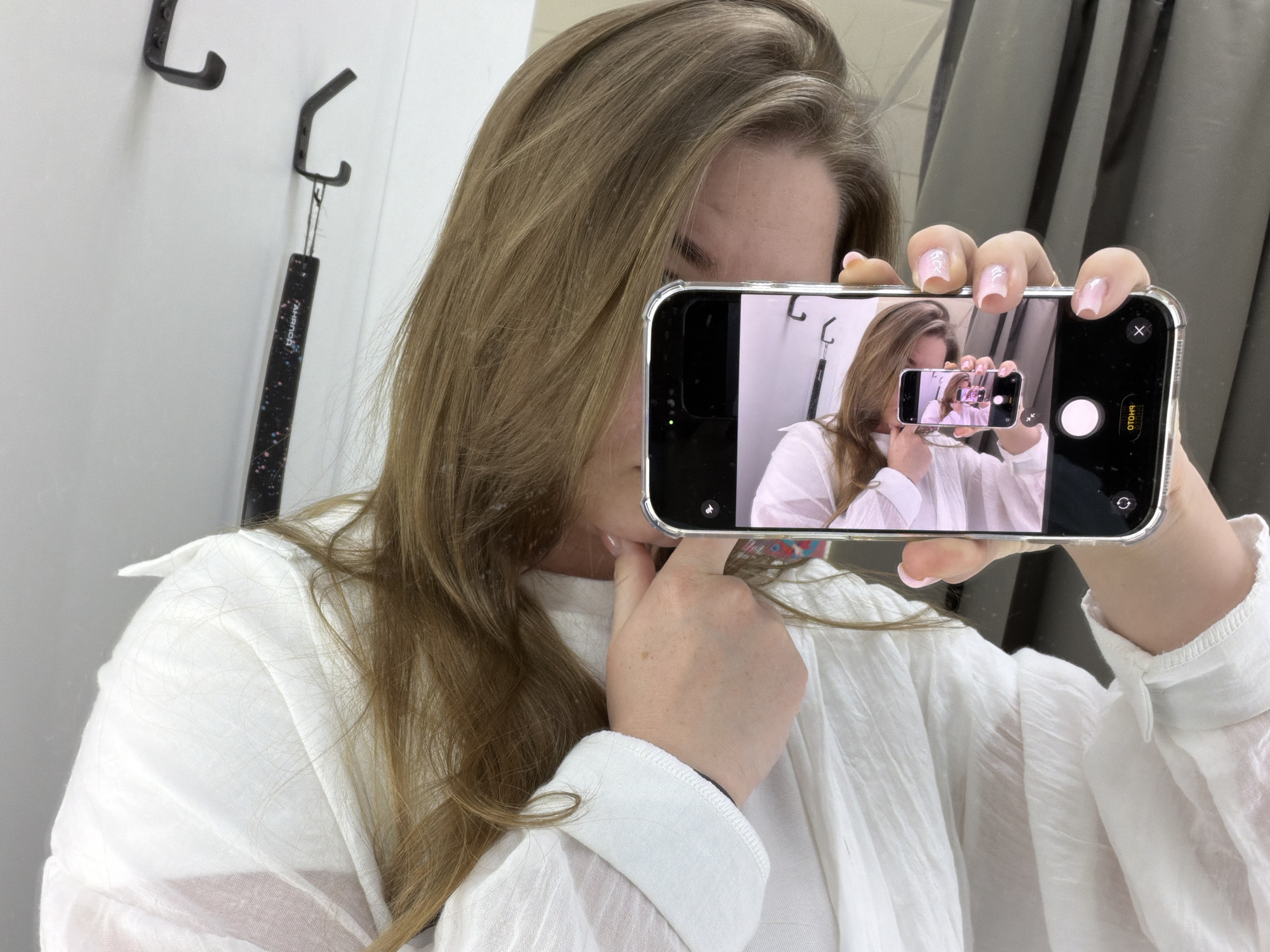 A woman with long brown hair is taking a selfie in what appears to be a dressing room or similar interior space. She is wearing a white collared shirt. She is holding up her smartphone, which is encased in a clear phone case, capturing her reflection as she takes the photo. On the phone's screen, we see a smaller version of herself, also taking a picture with her phone. In the background of the scene, hooks are visible on a white wall. The location is identified as Chaikovsky, Russia.