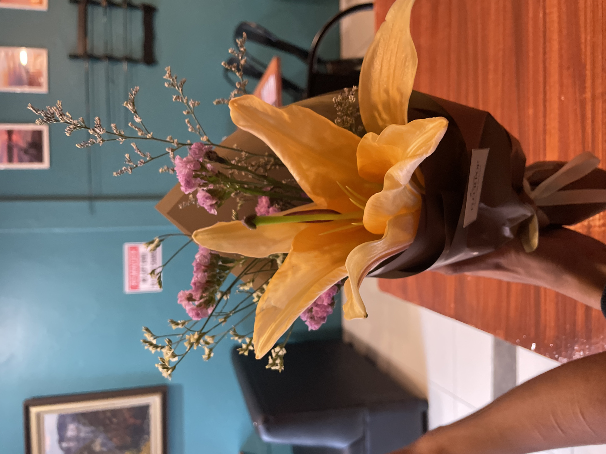 The image shows a close-up of a hand holding a bouquet of flowers. The bouquet contains a large orange lily, surrounded by smaller pink and white flowers and delicate dried foliage. The flowers are wrapped in dark brown paper and tied with a white ribbon. A label on the paper reads "FLORA TOP." The background suggests an indoor setting, possibly a cafe or restaurant, with a teal-colored wall, framed pictures, a dark leather chair, and a wooden table. The location is identified as Davao City, Philippines. The overall lighting is warm and soft, creating an inviting atmosphere.