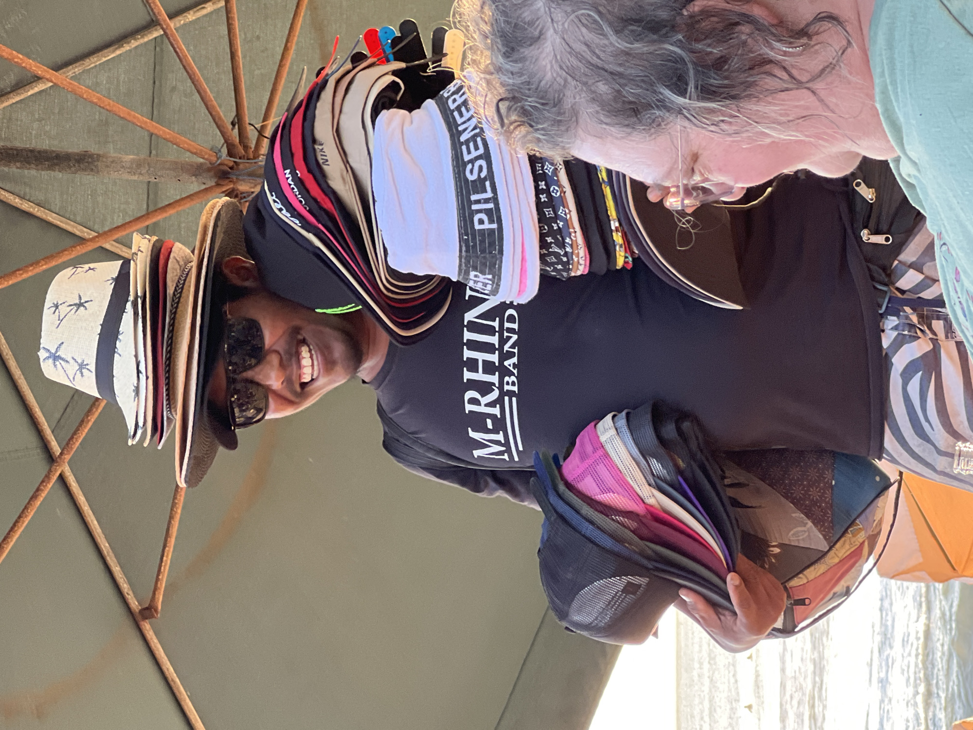 In Kuta, Indonesia, a man stands under a large umbrella, selling hats. He wears sunglasses and a black t-shirt with white lettering that reads "M-RHINE BAND." He holds a stack of baseball caps in his left arm and a tall tower of various hats, including straw hats and bucket hats, in his right hand. A woman with gray curly hair and glasses is visible in the foreground, looking at the man. The background shows a beach with the ocean and a few people in the water.