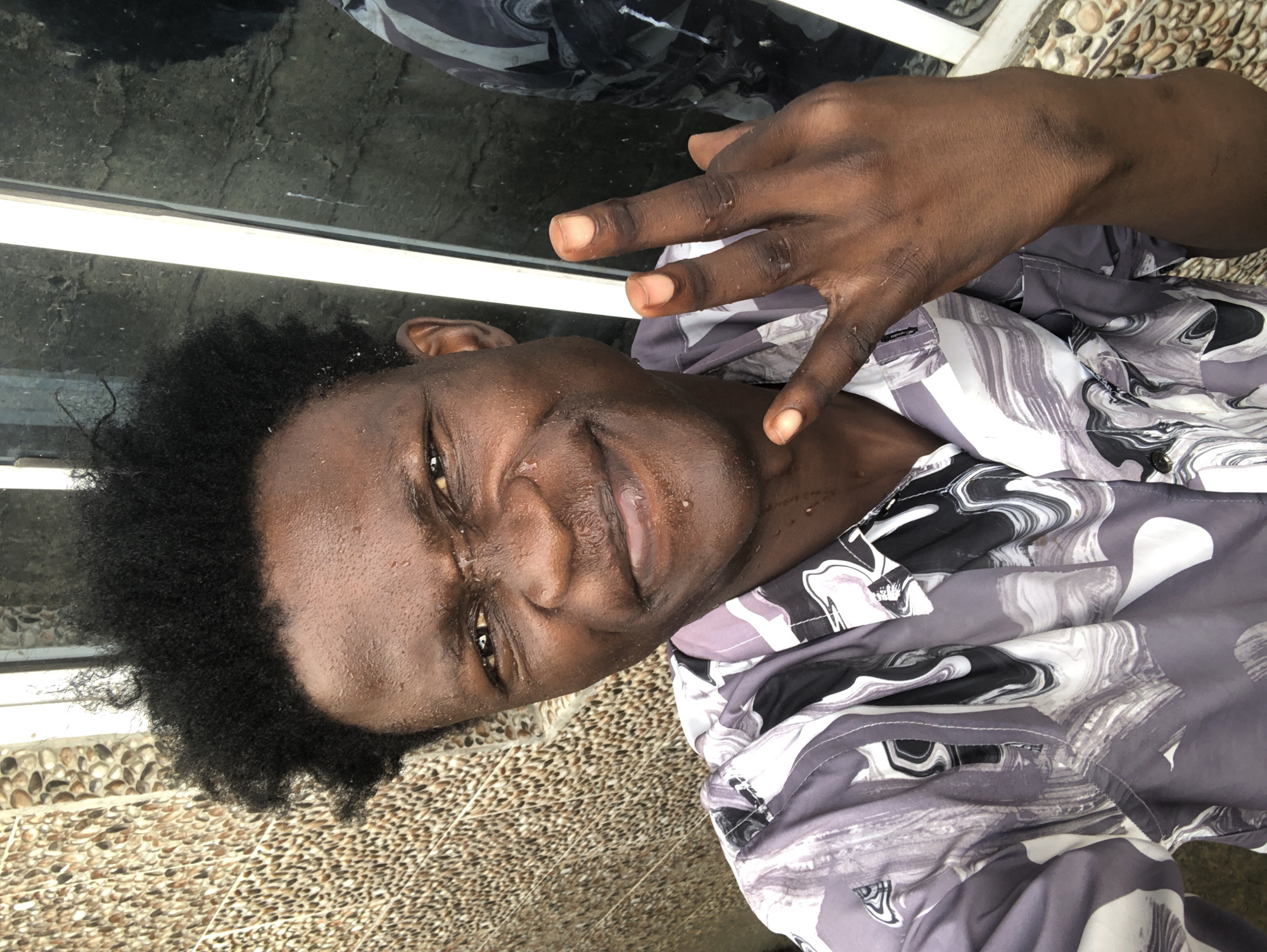 A dark-skinned male, with short, curly black hair, is captured in a close-up selfie. He is wearing a gray and white patterned button-up shirt. Water droplets are visible on his face and neck, indicating he may be sweating or has recently been in water. He is making a hand gesture with his right hand, with his index and middle fingers extended upwards. The background shows a textured wall and a window. The scene appears to be outdoors in a residential area.