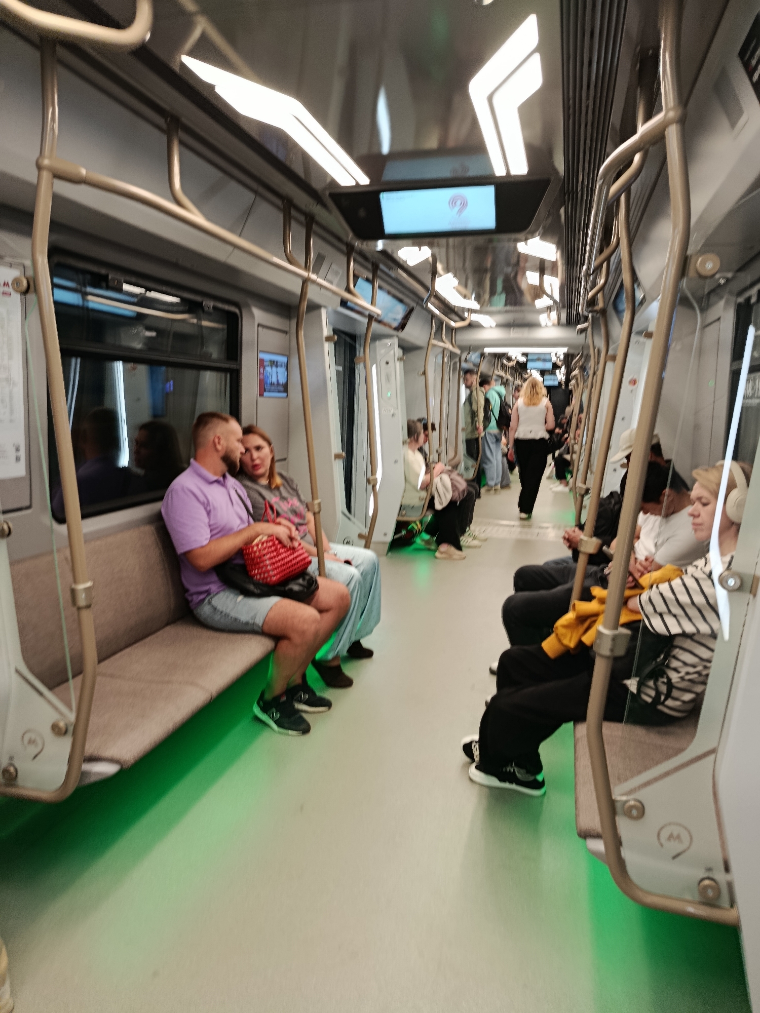 The image shows the interior of a modern subway train. In the foreground on the left, a man wearing a purple polo shirt and denim shorts sits with a woman wearing a gray t-shirt and light blue pants. The woman holds a red woven bag on her lap. They are seated on a beige bench that runs along the side of the train car. Green light emanates from beneath the bench, illuminating the floor. Several other passengers are visible further down the car, some seated and some standing. The train car features metal grab bars and overhead handrails, along with large windows. A digital display screen is visible on the ceiling of the train. The overall impression is that of an active public transportation environment.