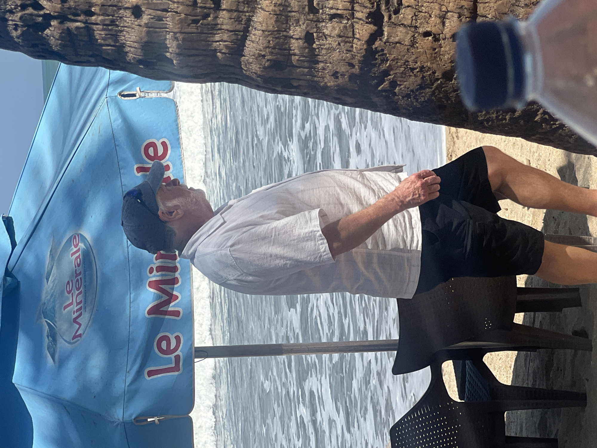 A middle-aged Caucasian man with a white short-sleeved button-up shirt, black shorts, and a dark baseball cap stands on a sandy beach in Kuta, Indonesia, facing to the right. He is partially obscured by a palm tree trunk on the right side of the frame and a large blue umbrella with "Le Minerale" branding on the left. The ocean with gentle waves is visible in the background, with white foam and blue water. Two dark plastic chairs are positioned in the sand in front of the man. A plastic water bottle with a blue cap is in the foreground, slightly out of focus. The lighting suggests a sunny day.