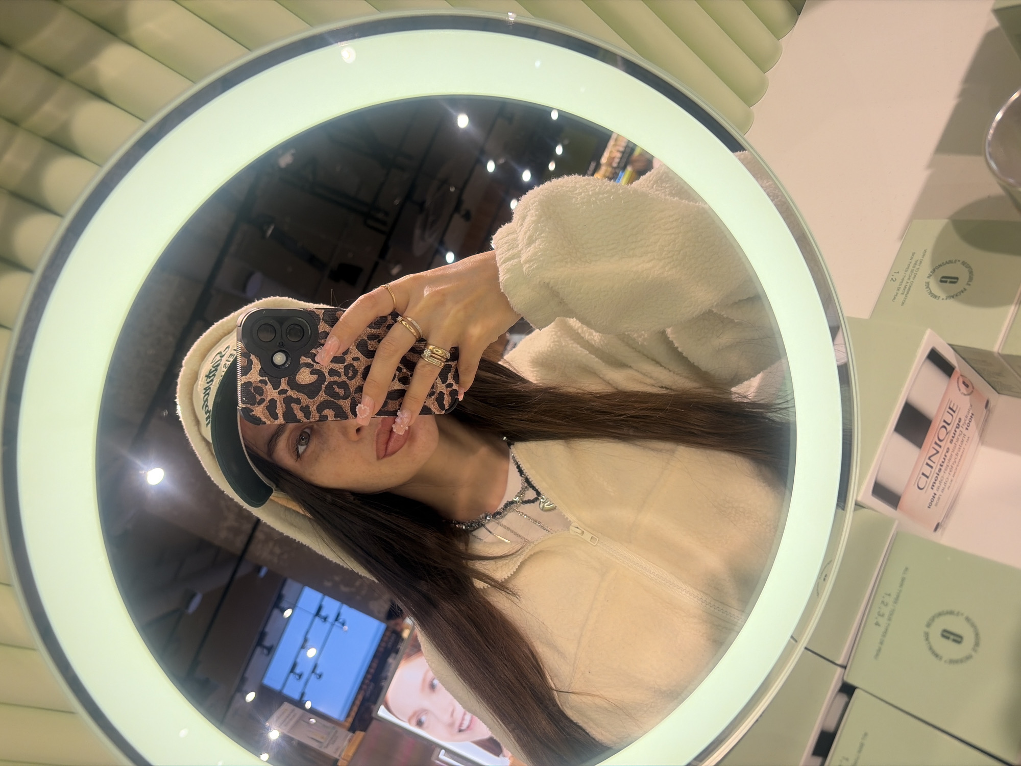 A young woman with long, dark hair is taking a mirror selfie. She is wearing a cream-colored fuzzy zip-up jacket and a green and cream baseball cap. She is also wearing a necklace with a silver heart pendant and multiple rings on her fingers. Her phone, with a leopard-print case, is held up to take the photo. In the reflection of the mirror, a bright ring light is visible around the edge. In the background, there are shelves with boxes of Clinique products, suggesting the location is a cosmetics store. The overall environment appears to be a brightly lit retail space. The specific location of Saint Petersburg, Russia is not discernible from the image.