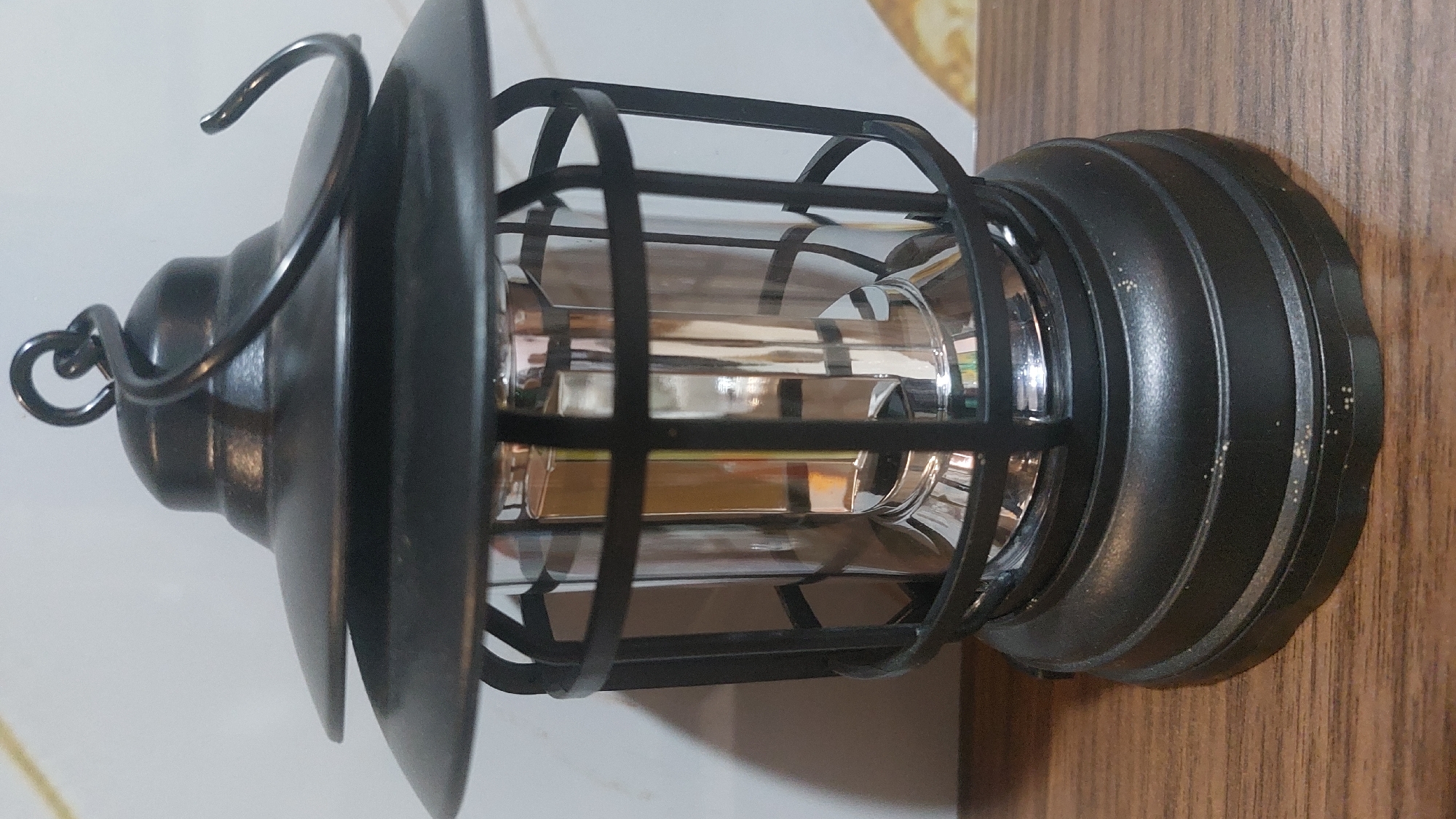 A close-up, eye-level shot showcases a black, vintage-style LED lantern sitting on a wooden surface. The lantern has a tiered, conical top with a decorative hook. A black metal cage surrounds a clear, cylindrical glass housing, which appears to contain a reflective, metallic element, likely the LED bulb and its mechanism. The base of the lantern is also black and has ribbed detailing. The background is a plain, light-colored wall. The image is lit from the front, casting a slight shadow behind the lantern. The overall impression is of a decorative or functional item, possibly placed indoors. There are no people or animals present. The image is crisp and in focus, with the texture of the wood and the metallic sheen of the lantern clearly visible. There is no indication of the location being the City of Medan, Indonesia; the scene appears to be a generic indoor setting.