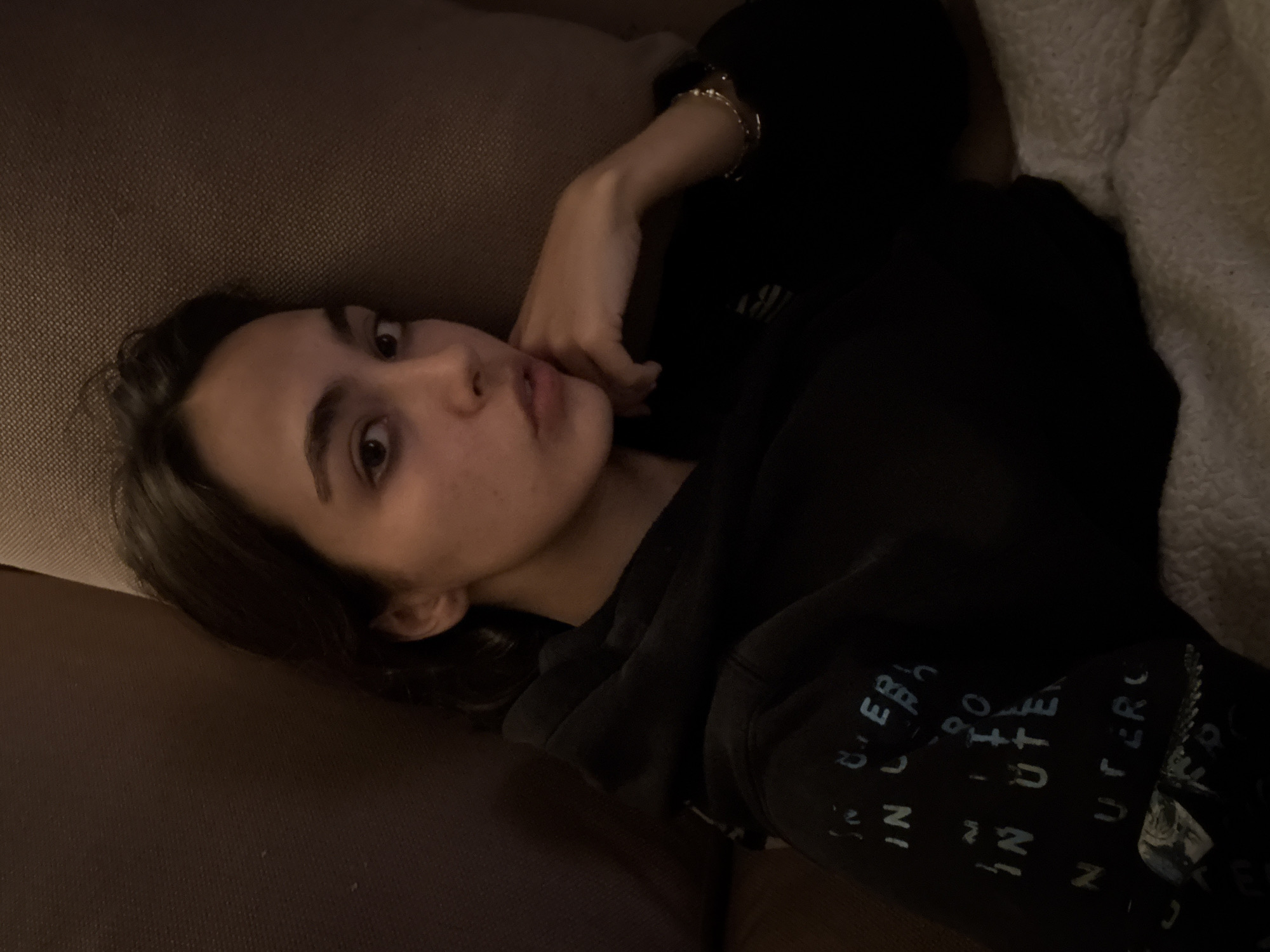 A person is lying on a brown couch with a fluffy white blanket to their right. They are wearing a black hooded sweatshirt with white writing on the sleeve. The person's face is visible, and they have brown hair and brown eyes. They are looking at the camera. The location appears to be indoors, and the lighting is dim. No information suggests this photo was taken in Mexico City, Mexico.