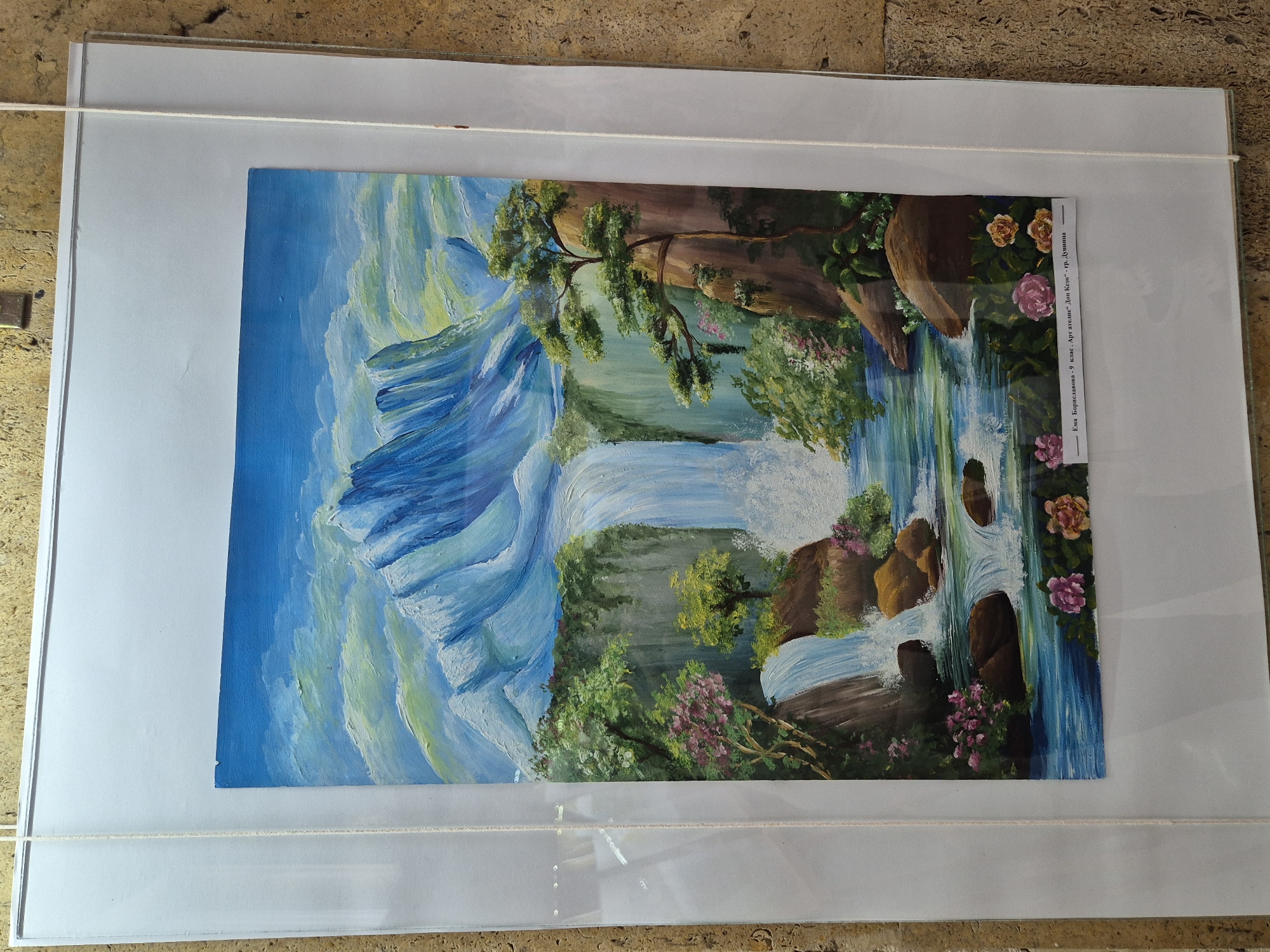 A framed painting of a mountain landscape with waterfalls hangs indoors. The painting features stylized blue and white mountains on the left, flowing into waterfalls cascading through rocky cliffs with lush vegetation and flowers on the right. A small label is affixed to the right side of the painting, containing text in a language that is not discernible in the provided image. The frame is white, and a white string hangs across the top of the frame. The background appears to be a textured surface. The location of Srebarna, Bulgaria, is not apparent.
