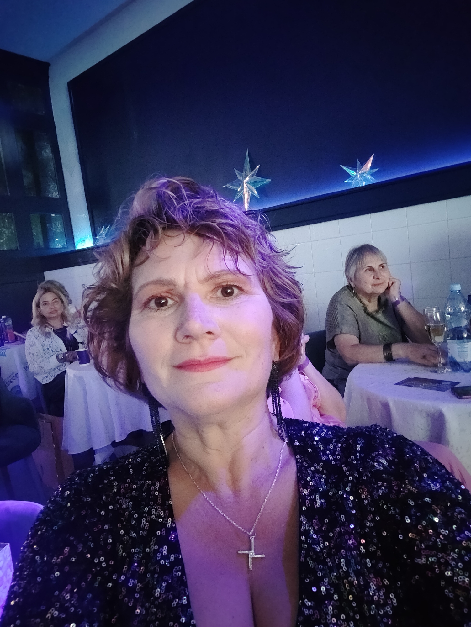 In a dimly lit venue with blue and purple lighting, a woman with short, curly brown hair and purple-tinted highlights takes a selfie. She is wearing a sparkly black dress and a silver cross necklace. Her eyes are looking directly at the camera, and her lips are slightly parted, with a hint of lipstick. In the background, two other women are seated at tables. The woman on the left has blonde hair and is wearing a white lace top. The woman on the right has grey hair and is wearing a green top, resting her chin on her hand. Two star-shaped decorations are visible on the dark blue wall behind them, and there are white tiled walls further back. Bottles of water and glasses of champagne are on the tables. The overall atmosphere suggests a social gathering or event. The location is not specified as Kyiv, Ukraine, in the image.