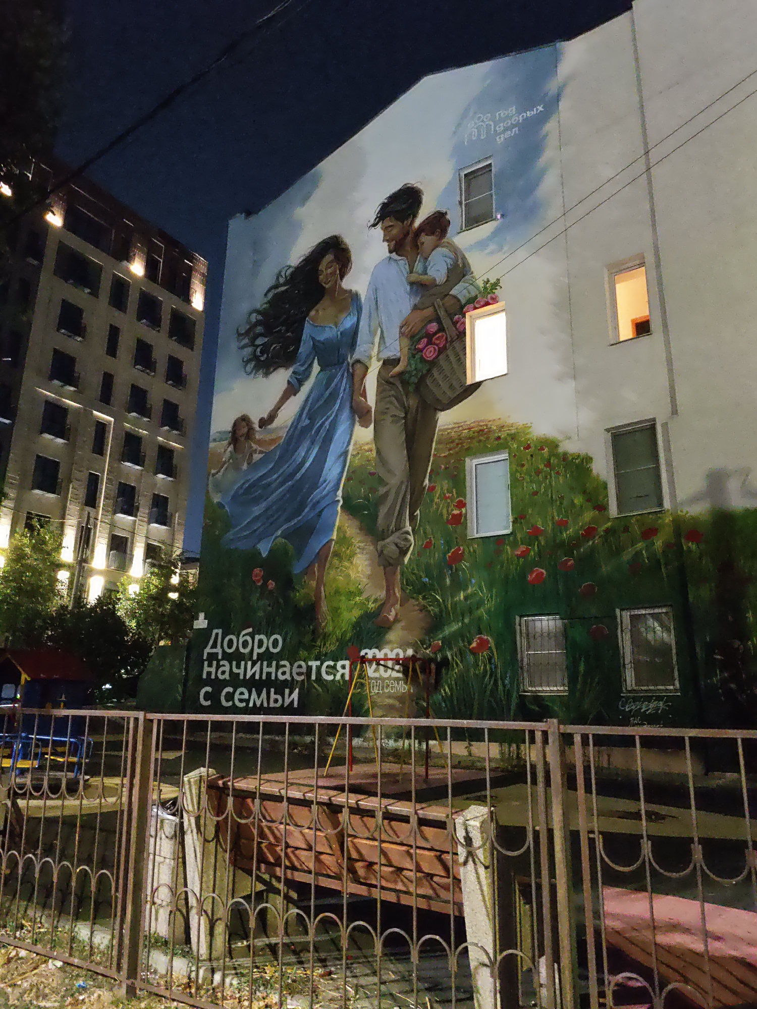 A mural on the side of a building depicts a family. A man carries a child on his shoulders, while a woman walks beside him, holding a basket of flowers. The mural is set against a backdrop of a field of red flowers and tall grass, with a cloudy sky above. Text in Russian reads "Добро начинается с семьи," which translates to "Goodness begins with family." The mural is located in Rostov-on-Don, Russia. In the foreground, a rusty metal fence surrounds a playground area with a slide and other equipment. The surrounding buildings are lit up, indicating it is nighttime.