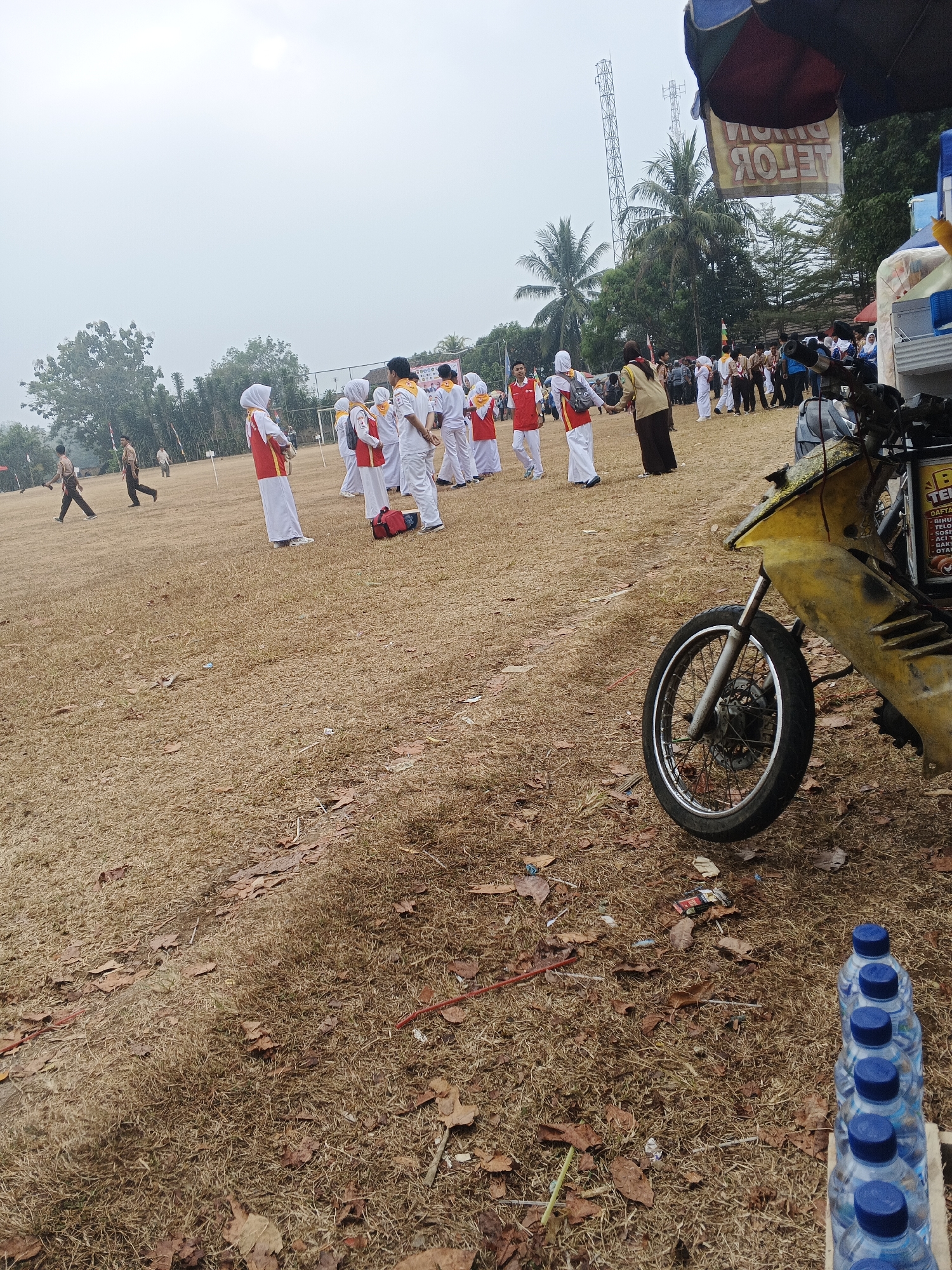 The image captures an outdoor scene, likely an event or gathering, in Warungkiara, Indonesia. The foreground is dominated by dry, brown grass littered with fallen leaves. To the right, several plastic bottles of water with blue caps are lined up on a wooden structure. Further into the scene, a yellow motorcycle is parked, its front wheel and part of the frame visible. In the background, a group of people, many wearing white uniforms with red vests or accents and some with yellow sashes, are gathered on the grassy field. Some individuals are wearing headscarves. They appear to be standing in groups or lines, possibly waiting for an activity or participating in a ceremony. A few other individuals in different attire, including a scout-like uniform, are also present on the field. In the far background, trees, power lines, and communication towers can be seen against a bright, overcast sky. A banner with text is visible hanging from an awning in the upper right portion of the image.