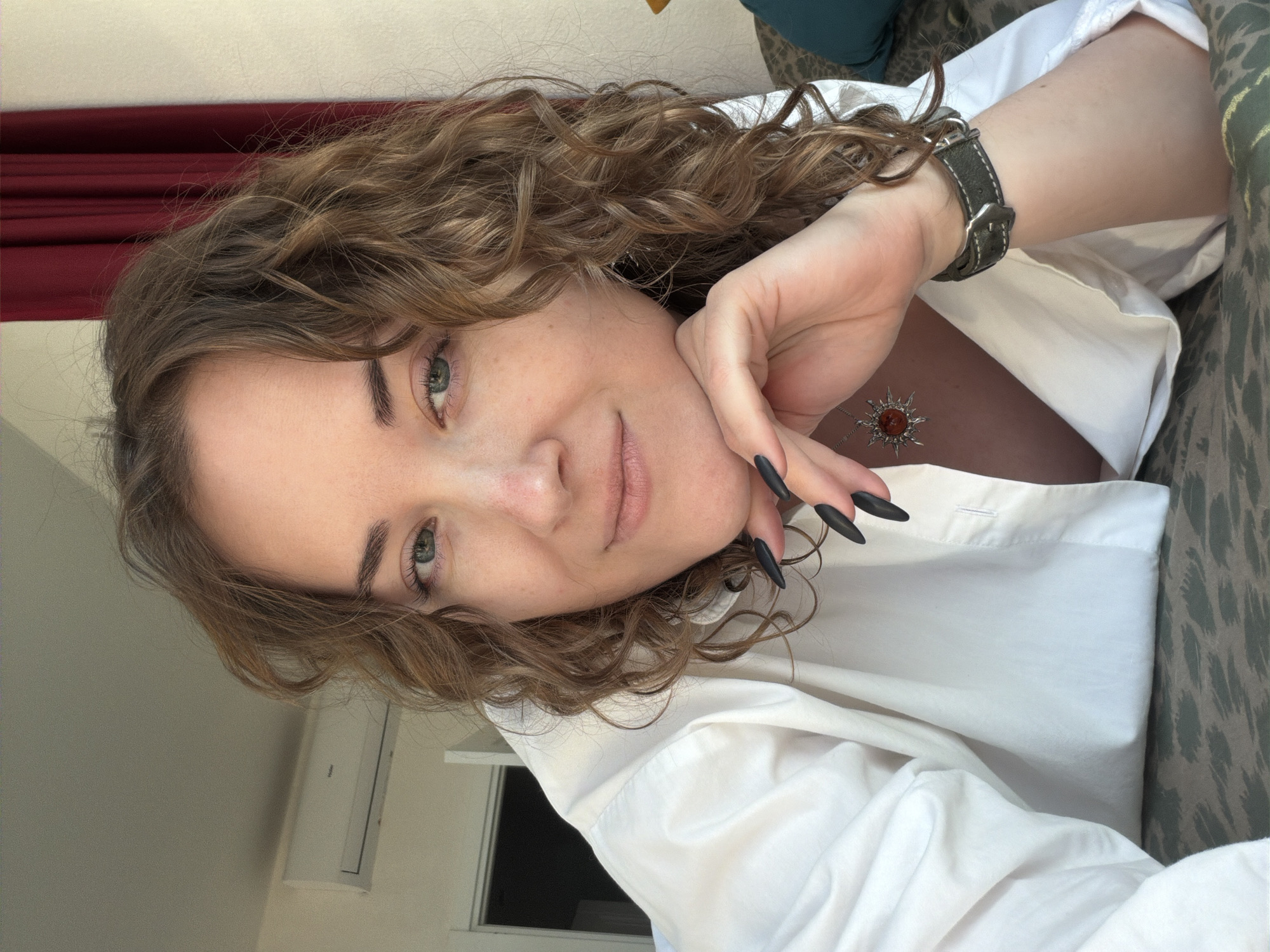 A woman with curly, shoulder-length hair poses for a selfie. She is wearing a white collared shirt and a watch with a dark leather strap. Her fingernails are long and painted black. She wears a pendant necklace with a sunburst-shaped pendant. The location is an indoor setting in the city of Unknown, Unknown. Red curtains are partially visible, and a window and air conditioning unit are in the background. The woman appears to be sitting or lying on a patterned couch.