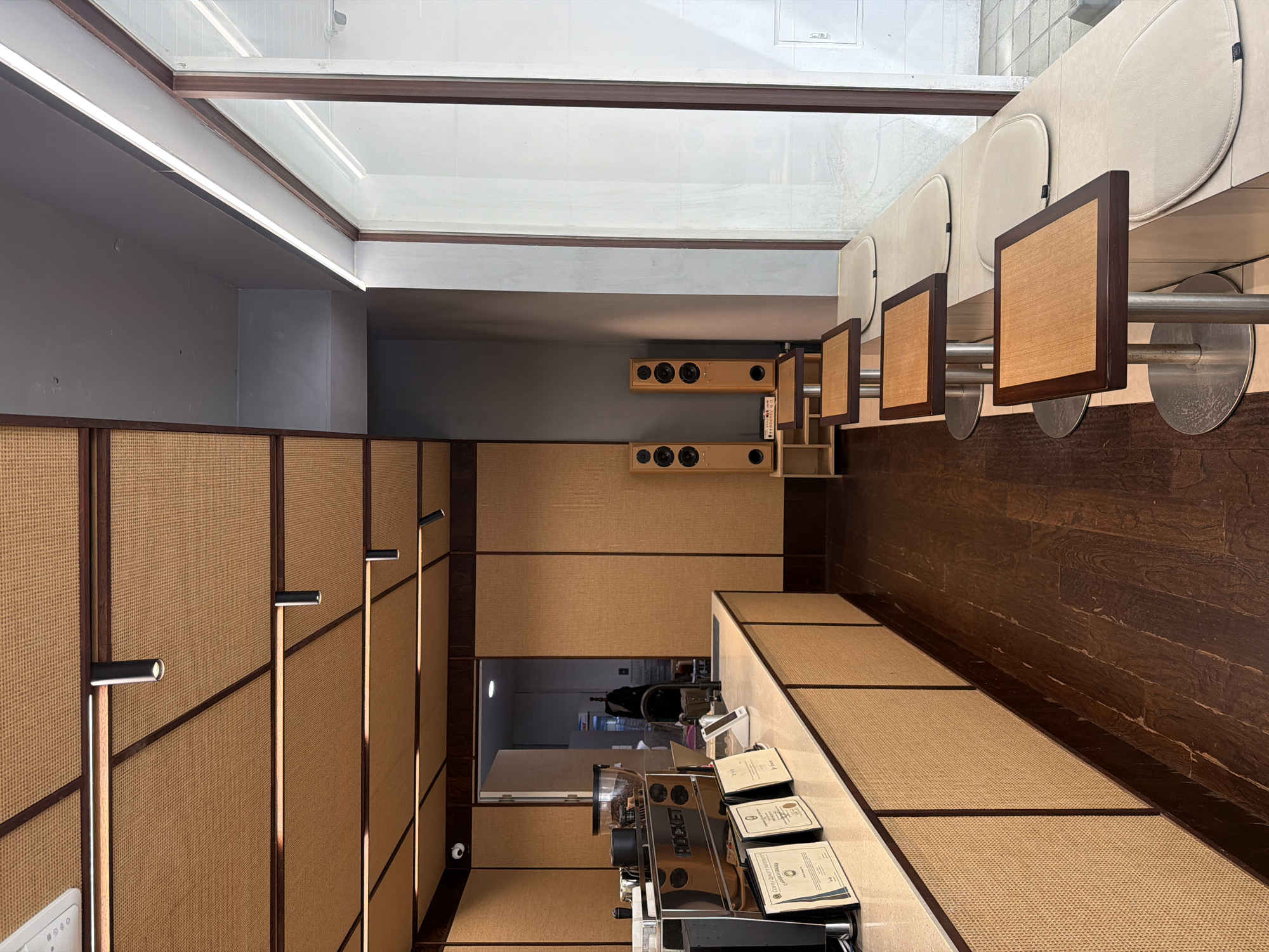 This appears to be an interior shot of a cafe located in Seoul, South Korea. The location seems meticulously designed, featuring a contemporary style with wooden panels and wicker-like wall coverings. The design elements include sleek, minimalist light fixtures and a row of speakers mounted on the wall. The seating includes a counter with backless chairs attached by poles. A modern espresso machine sits on the counter along with certificates. The room is lit by natural light coming from the ceiling window and wall fixtures. There are no people visible in the image.