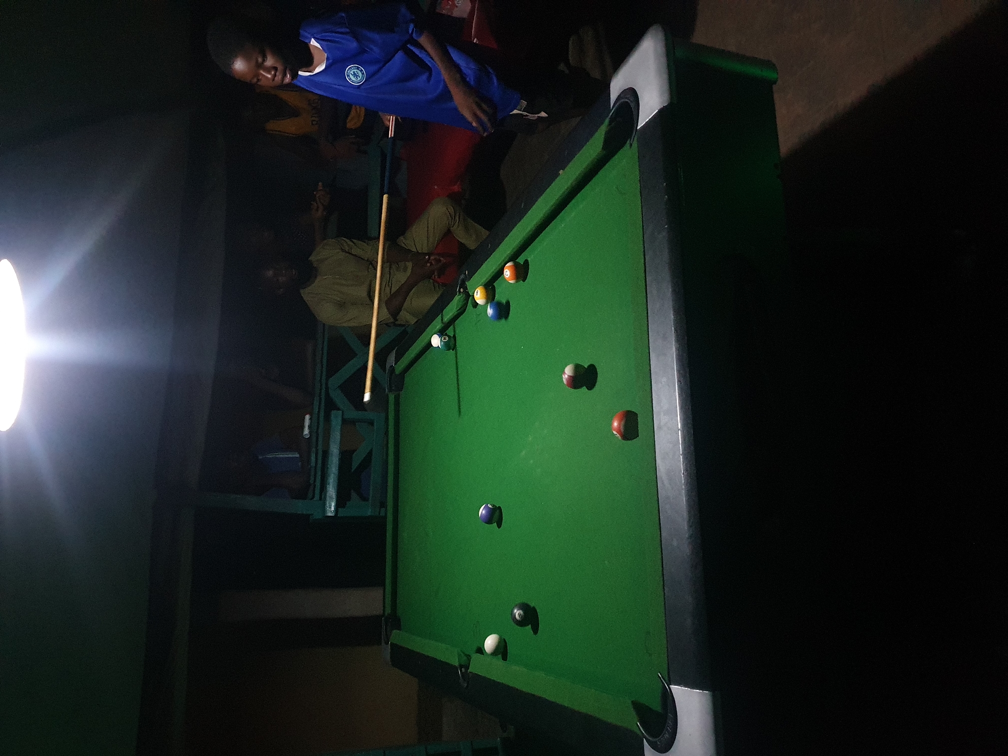 The image depicts an indoor scene at night, likely a recreation area, possibly in Lokoja, Nigeria, given the context. A green billiard table with several balls is centrally placed. A young boy in a blue t-shirt is standing to the right, holding a billiard cue, appearing to be playing. Another person, an adult male in a light-colored garment, is seated behind the table, also holding a cue. In the background, other individuals are partially visible, seated on benches or standing. The lighting is dim, with a bright overhead lamp illuminating the table and players. The environment appears to be a rustic, possibly open-air structure, with wooden elements and a dark background suggesting night time. The overall atmosphere suggests a casual gathering for a game of billiards.