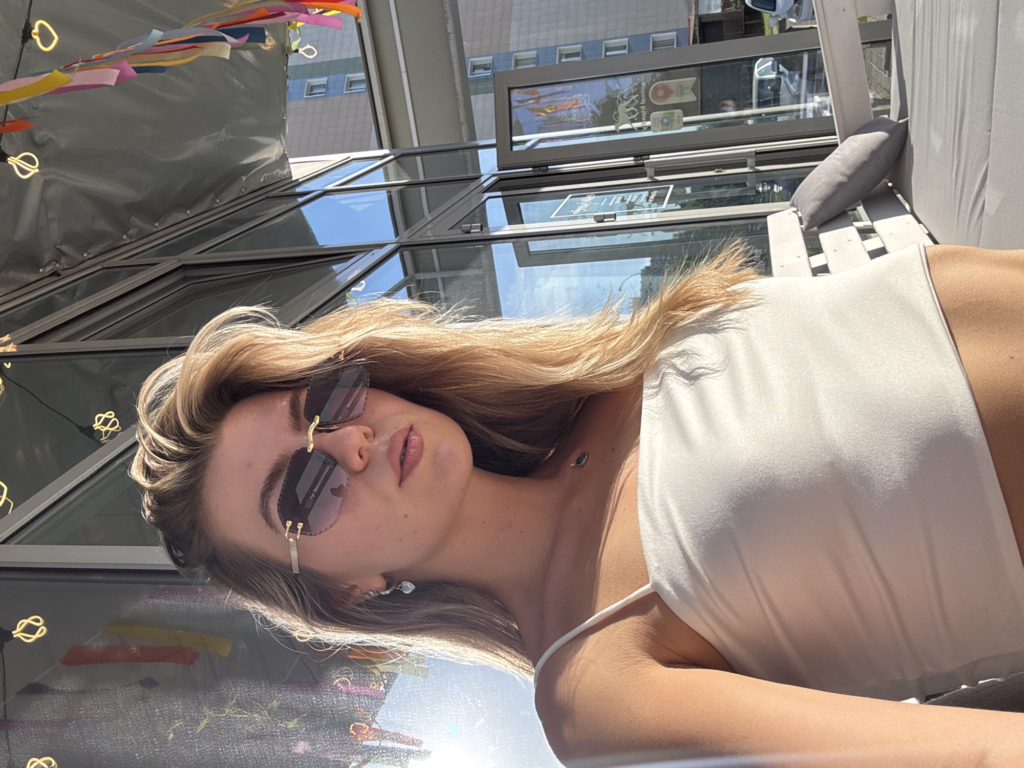 A woman wearing a white top and sunglasses is reclining on a bench or chair in an outdoor setting. She is facing the camera with a slight tilt. Above her is a glass or transparent structure, with buildings visible in the background. Colorful decorations hang from the ceiling above the structure. The location appears to be an urban or semi-urban setting.