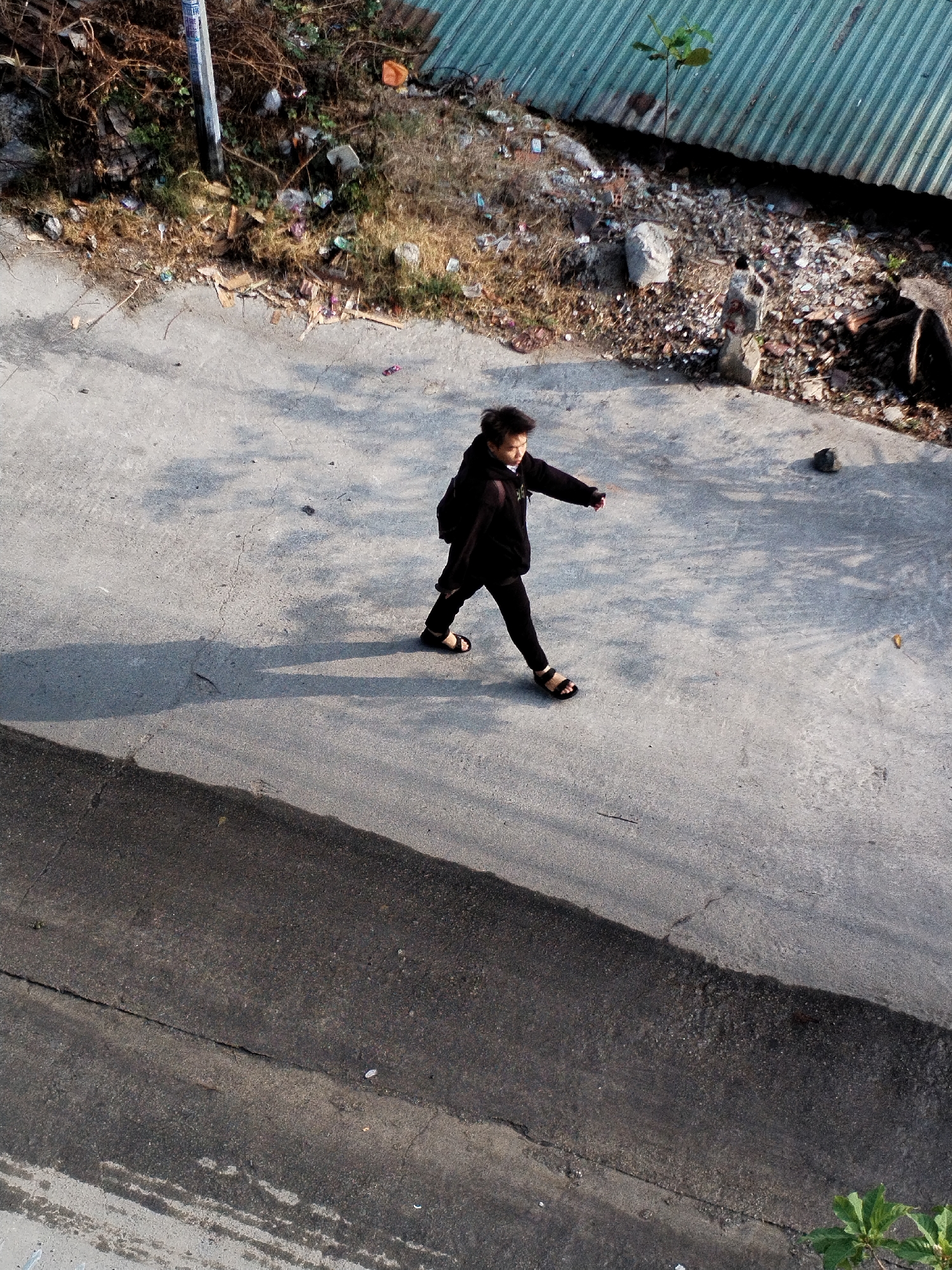 A person wearing a black sweatshirt, black pants, and sandals is walking on a concrete surface, likely a sidewalk or a paved area, in an outdoor environment. The person has dark hair. The area to the top left is a patch of vegetation, with a variety of small plants and debris. To the right, a corrugated metal fence or wall is visible, with some more vegetation and debris in front. A road of darker asphalt is to the bottom, and to the center right, is what is possibly an elevated sidewalk area. Nha Trang Ward, Vietnam can not be confirmed from this image alone.