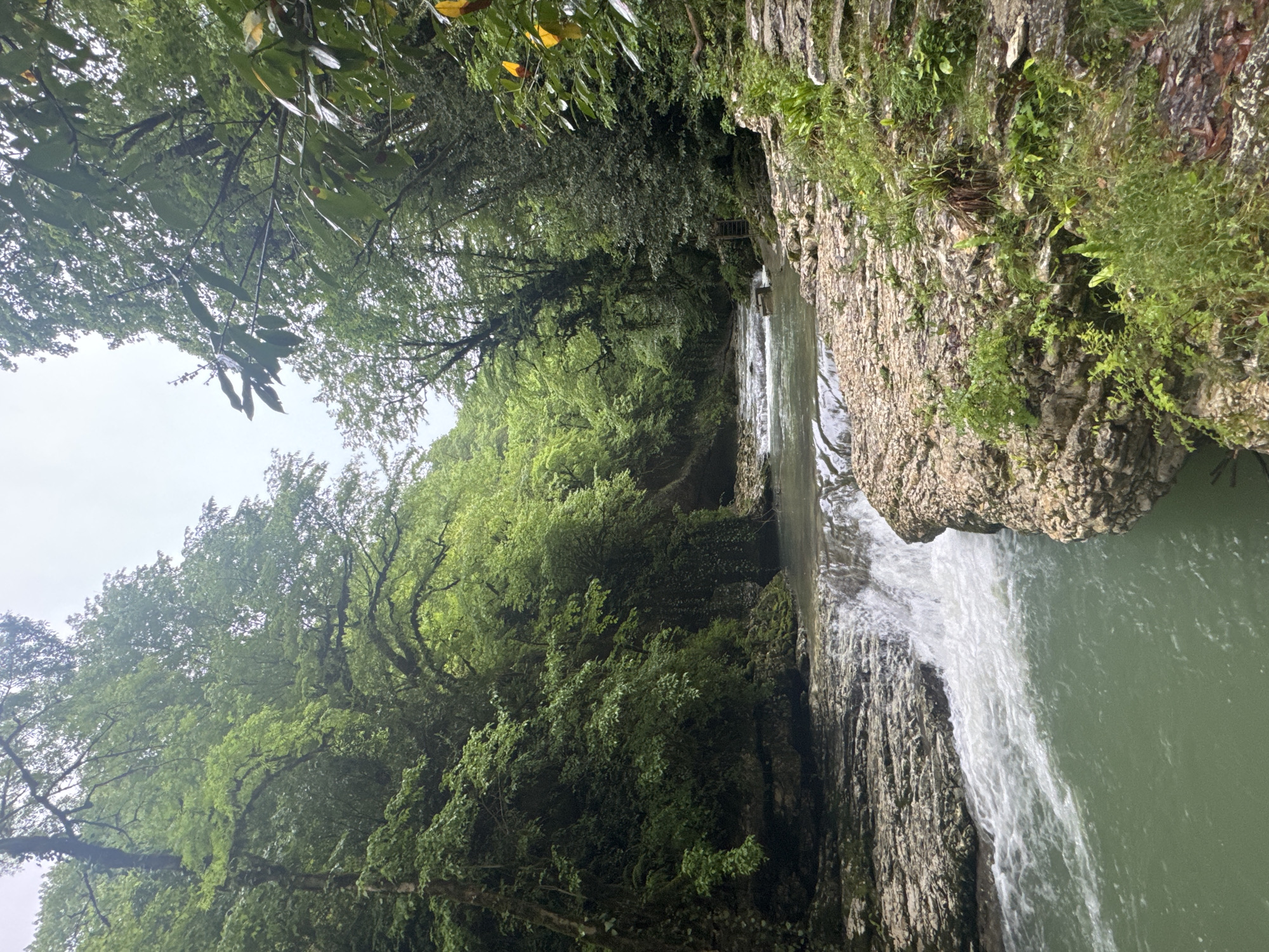 The image depicts a waterfall in Tsabal, Abkhazia, surrounded by lush greenery. Water cascades over a rocky cliff face into a pool below. The scene is framed by trees and foliage, indicating a natural, outdoor environment. The sky is overcast, suggesting cloudy weather conditions. No people are visible.