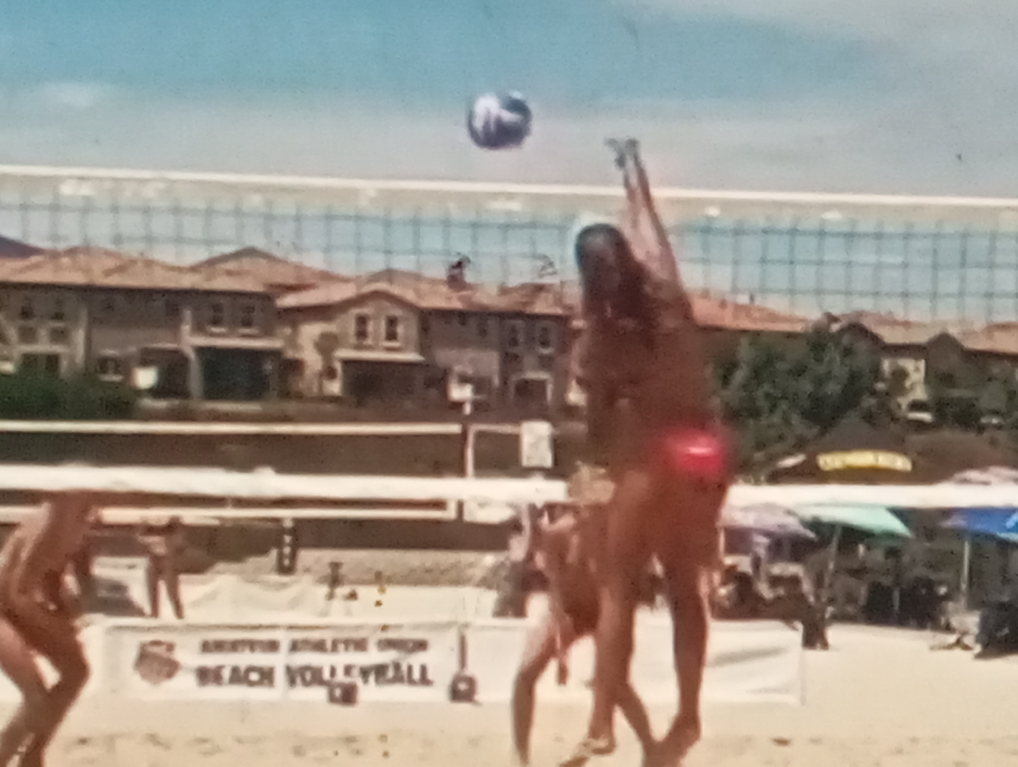 The image depicts a beach volleyball game. In the foreground, two women are playing on a sand court, with one in the process of hitting the ball. A volleyball is visible in mid-air above the net. The background shows houses, a volleyball net, and beach umbrellas. The overall scene suggests a sunny day at a beach venue. This image does not contain any information that would place it in Sofiivska Borshchahivka, Ukraine.
