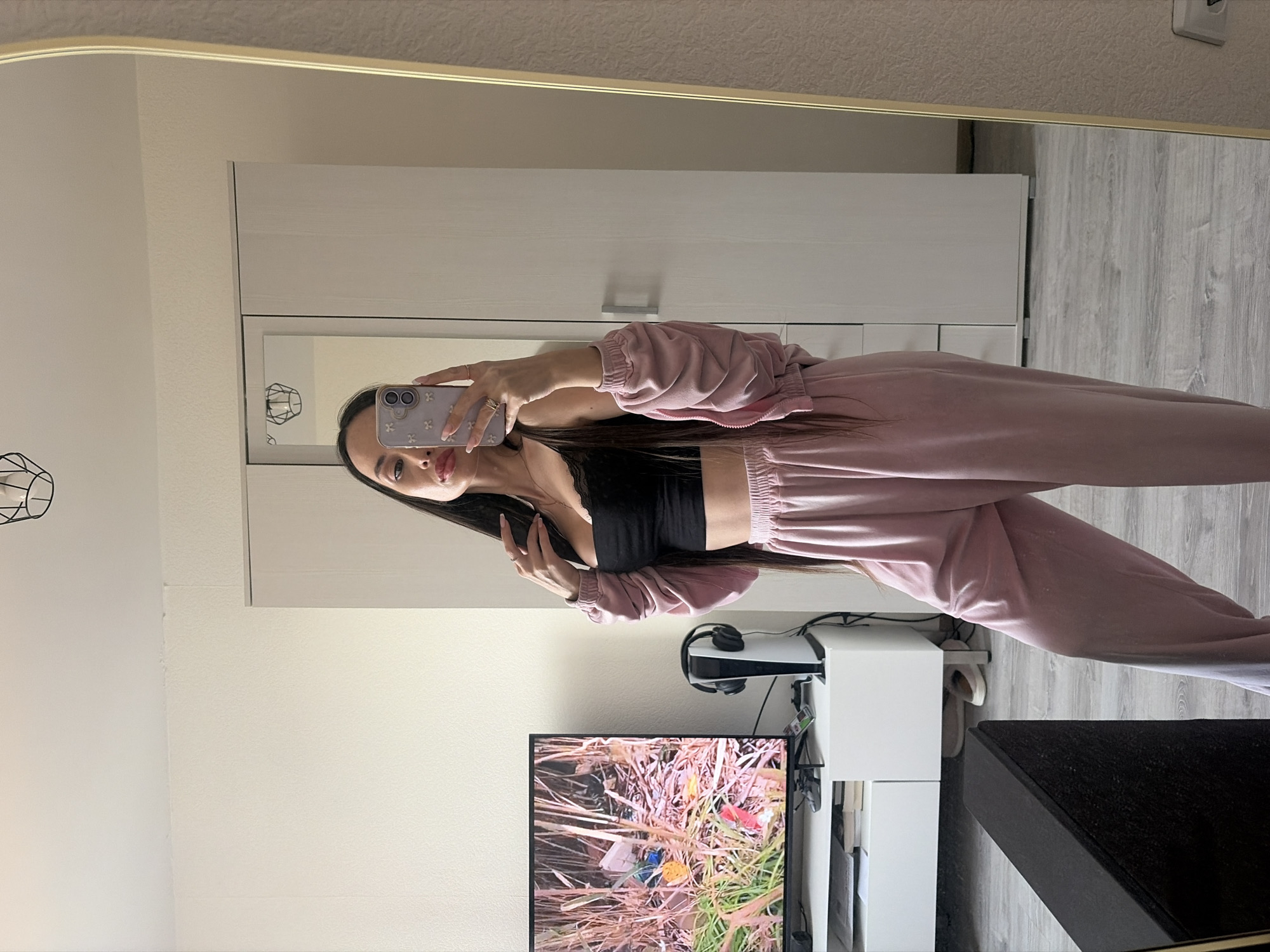 A woman is taking a selfie in a mirror inside a room. She is wearing a pink tracksuit and a black top. In the background, there is a white wardrobe, a TV showing a nature scene, a white TV stand, a PlayStation 5, and a black sofa. The walls are white, and the floor appears to be gray laminate. There's no direct evidence to confirm the location is Saint Petersburg, Russia.