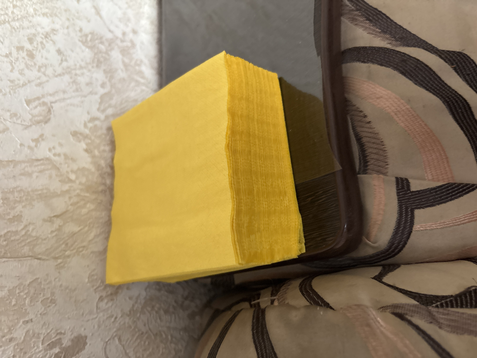 This image depicts an indoor scene in Chernogorsk, Russia. A substantial stack of bright yellow, square-folded napkins, estimated to be 20-30 units, rests on a dark, rectangular, and mildly reflective surface, likely a tray or tabletop. To the left, a section of upholstered furniture is visible, featuring a beige fabric adorned with abstract patterns of dark brown and light pink curvilinear stripes. The background consists of a light-colored wall exhibiting a heavily textured and uneven finish. No individuals or dynamic actions are present in the frame.