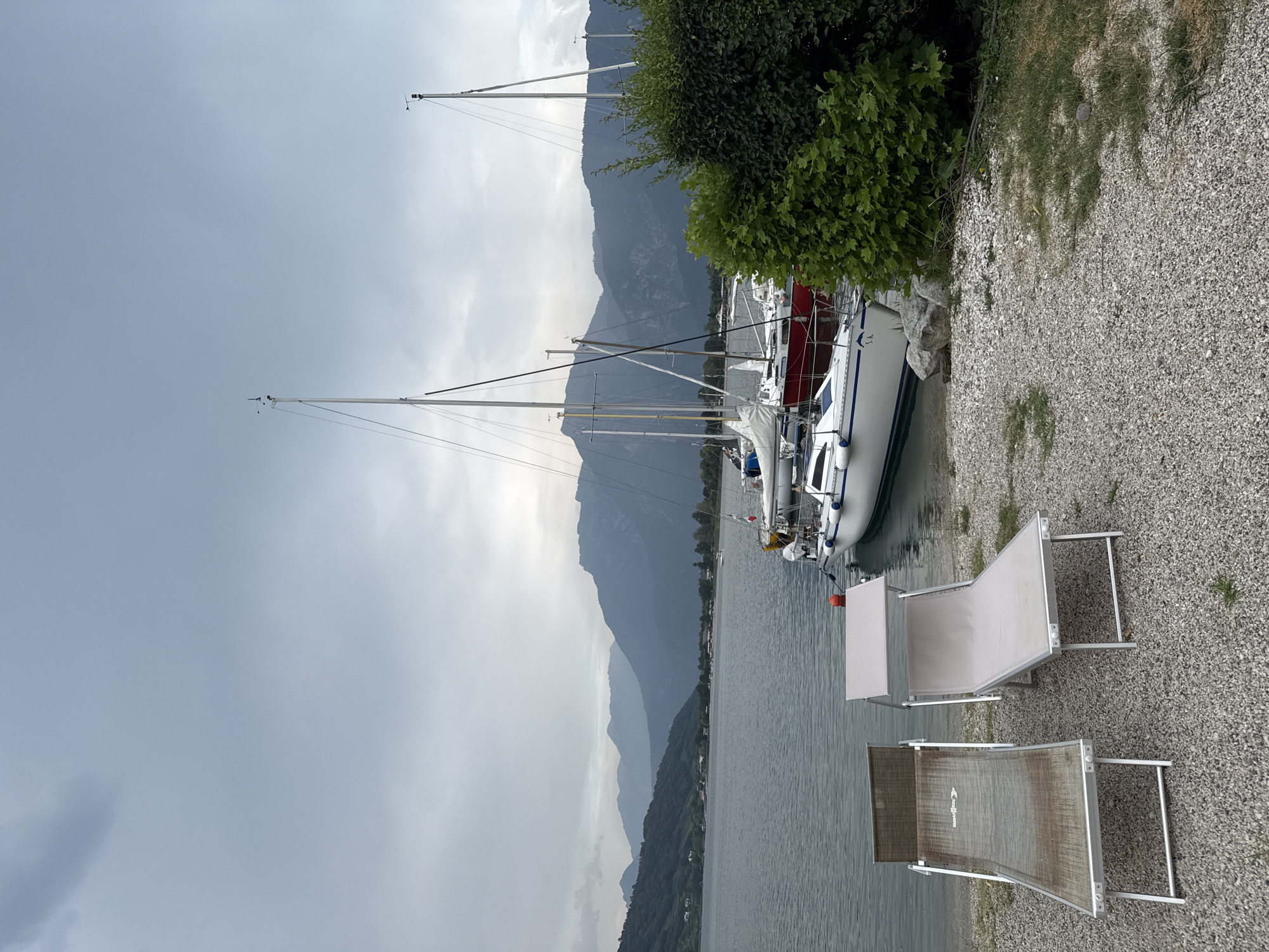 Two empty lounge chairs are positioned on a gravelly shore of a lake. In the background, several sailboats are docked, with their masts reaching towards a cloudy, overcast sky. Mountains form the backdrop across the lake. The scene appears to be a quiet marina area, possibly in or near Calceranica al Lago, Italy, given the prominent lake and mountain landscape.