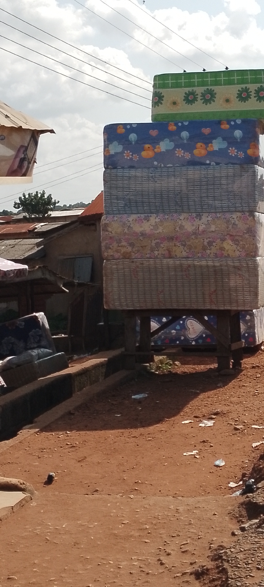 The image depicts an outdoor scene in Adikpo, Nigeria. In the foreground, a reddish-brown dirt ground is visible, with scattered debris including white plastic. To the left, a raised concrete ledge runs along the bottom of the frame. Behind this ledge, on the left side of the image, are stacked mattresses. The mattresses have various patterns: the bottom one is covered in a woven-like material, the next has a floral pattern, then a blue mattress with yellow ducklings and hearts, and the top mattress features a green plaid pattern with yellow flowers. A mattress with a dark floral pattern is also leaning against a concrete structure to the left.
In the middle ground, several makeshift structures with corrugated metal roofs are visible. To the right of the mattresses, a wooden stand supports the bottom of the stacked mattresses, and under this stand, another mattress with a blue background and white hearts is visible. The background consists of more rooftops and some foliage. Overhead, power lines stretch across a sky with scattered clouds. The scene appears to be a marketplace or a residential area where mattresses are being sold or stored outdoors. There are no people clearly visible in the image.