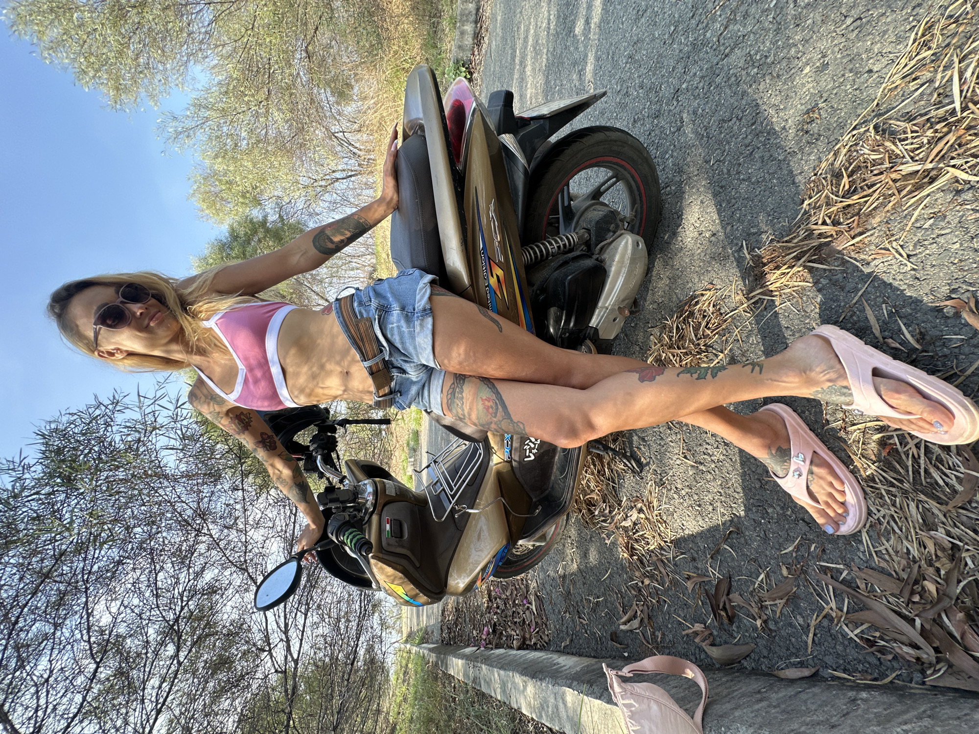 In Phu Thuy Ward, Vietnam, a woman with extensive tattoos on her arms and legs leans against a gold and brown scooter. She wears a pink sports bra, denim shorts, and pink flip-flops. The scooter, identified as a Honda PCX, is parked on an asphalt road covered with fallen leaves and dry grass. The background shows sparse trees and a clear blue sky, suggesting an outdoor, possibly rural or suburban, setting. A pale pink bag rests on a concrete curb to the left of the scooter. The woman is looking towards the camera with a relaxed posture.