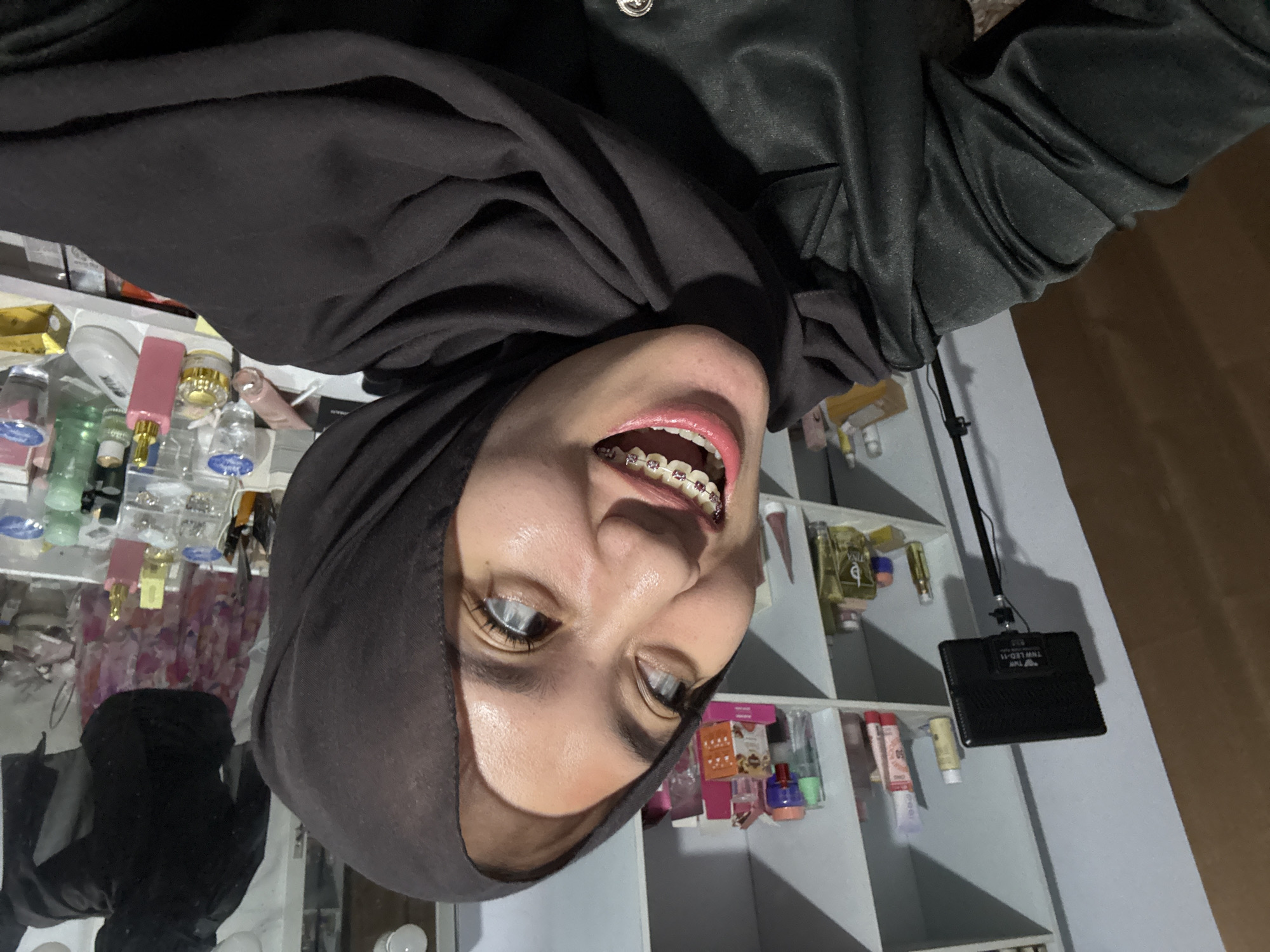 The media file is a vertically oriented, close-up, selfie-style photograph of a young woman wearing a black hijab and a black leather jacket. She is smiling broadly, revealing braces on her teeth and bright pink lipstick. Her eyes are wide open, and her eyebrows are well-defined.
The background consists of shelves filled with various cosmetic products, including bottles, tubes, and boxes of skincare and makeup items. Some of the products have colorful packaging. The shelves appear to be in a well-lit room, possibly a bedroom or a beauty store.
The lighting suggests it is indoors, and the time of day is not discernible from the image. The woman's expression is joyful and confident. There are no discernible location cues in the image that specifically point to Gadingrejo, Indonesia, beyond the provided context. The overall impression is a personal moment captured in a private space, showcasing the woman's smile and her collection of beauty products.