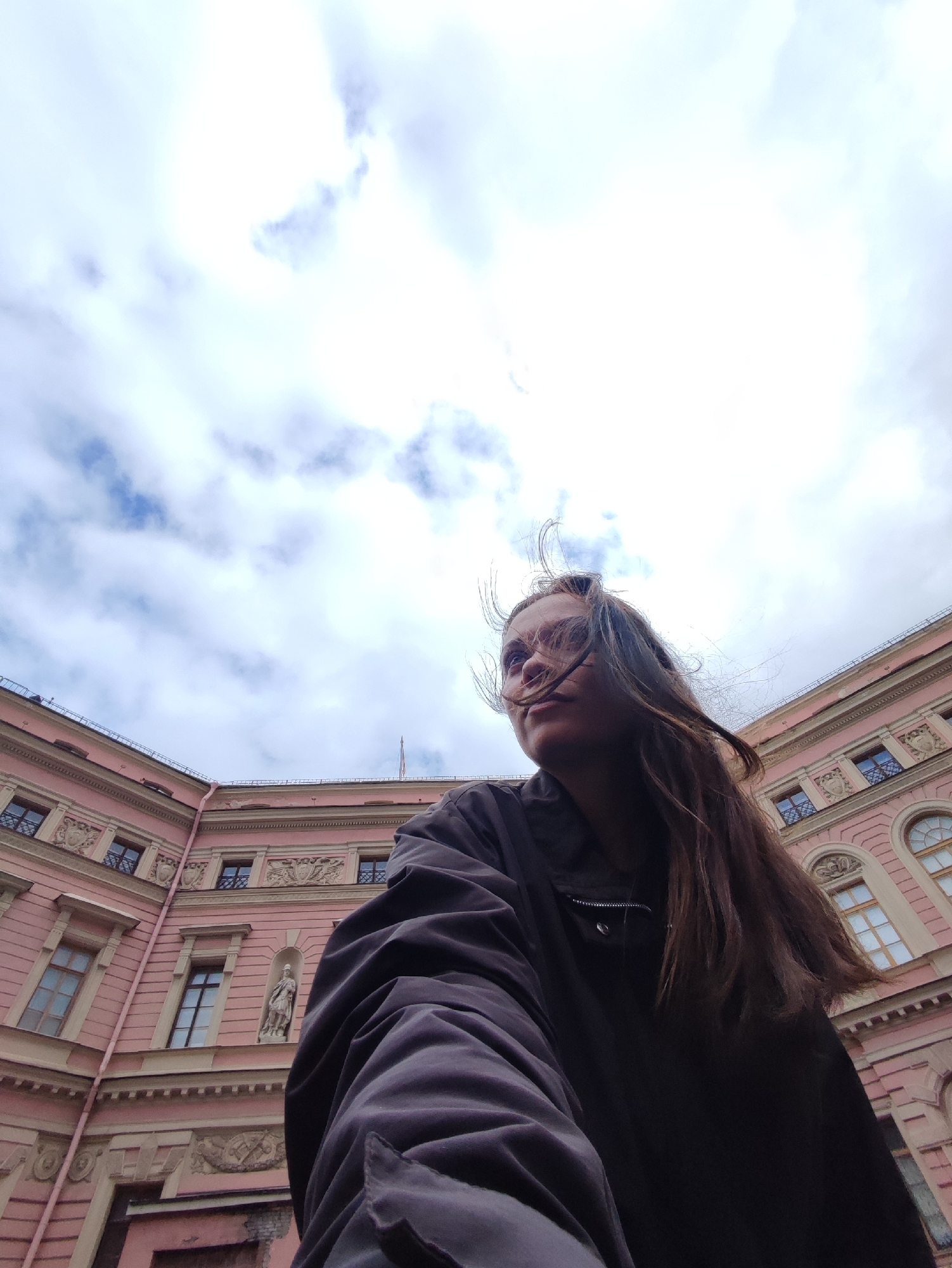 A low-angle, wide shot captures a young person with long, brown hair against a cloudy sky and a building with pale pink walls in Saint Petersburg, Russia. The person is wearing a dark jacket and is positioned in the foreground, with their face turned to the right. Their hair is blown across their face by the wind, obscuring part of their features. The building, which appears to be an ornate structure with multiple windows and decorative architectural elements, occupies the background and the right side of the frame. The sky is a mix of white and light blue, with visible cloud formations. The perspective suggests the photo was taken from a ground-level viewpoint, looking upwards.