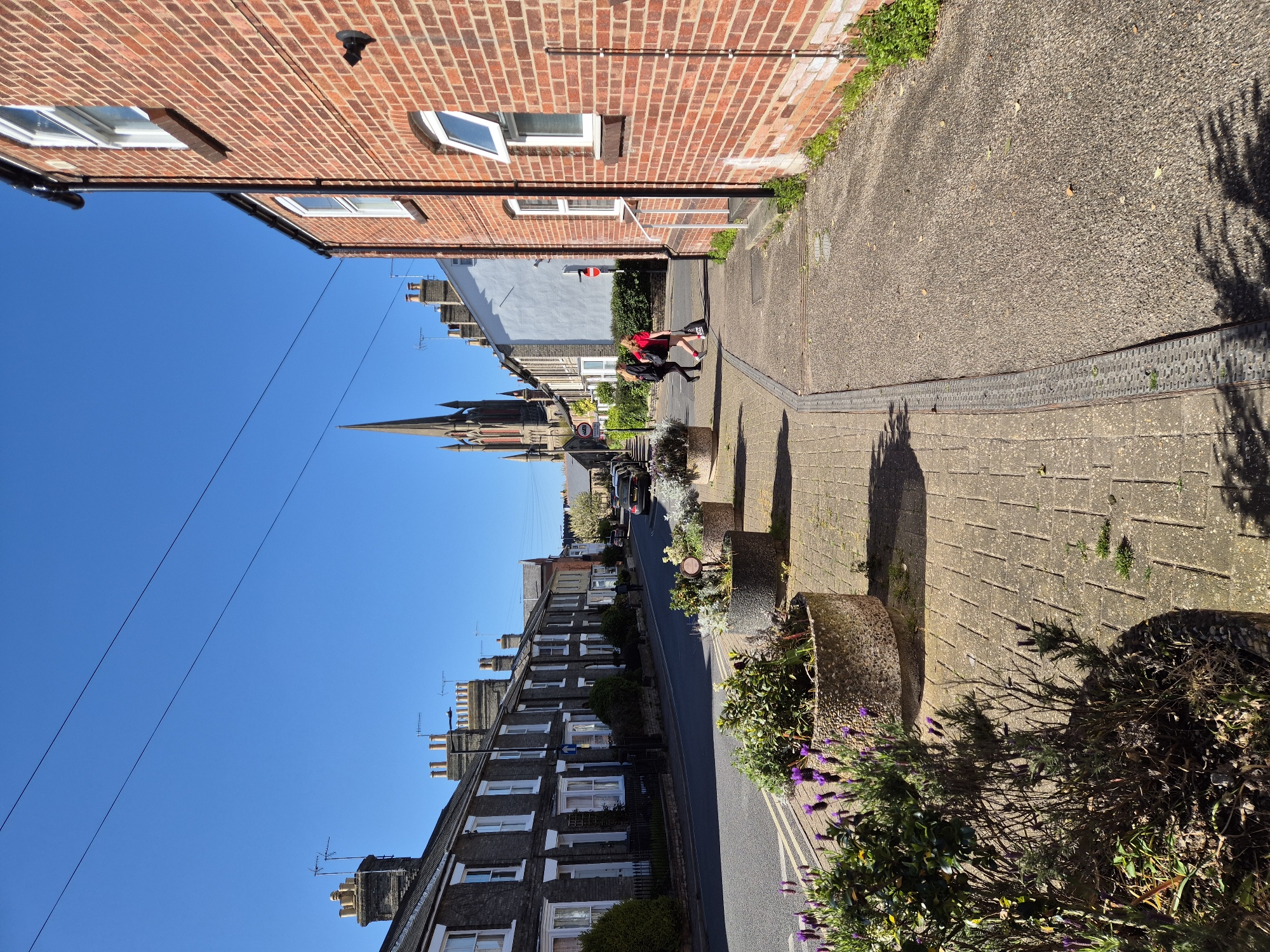 The image presents a sunlit street scene, likely a residential area in the UK, consistent with West Suffolk. The view looks down a street lined with buildings under a clear blue sky.
In the foreground on the right, the side of a red-brick building with several white-framed windows is prominent. Below it, a paved sidewalk features two large, weathered planters filled with greenery and purple-flowering plants. Long shadows are cast across this pavement, suggesting strong sunlight from either morning or late afternoon. Two individuals are walking on this sidewalk further down: one, appearing to be a child or young person, wears a red top, dark bottom, and carries a backpack; the other, slightly behind, is dressed in dark clothing.
On the left side of the street, a continuous row of terraced houses with dark brick or stone exteriors and multiple chimneys stretches into the distance. Several cars are parked along this side of the road. Further down, a distinctive church spire with a clock face rises above the rooftops on the left.
The street itself is asphalt, with double yellow lines visible in the distance. Overhead, power lines traverse the bright, cloudless blue sky. A small, unreadable red circular sign is visible further down the street on the right side. No other discernible text is visible on any objects.