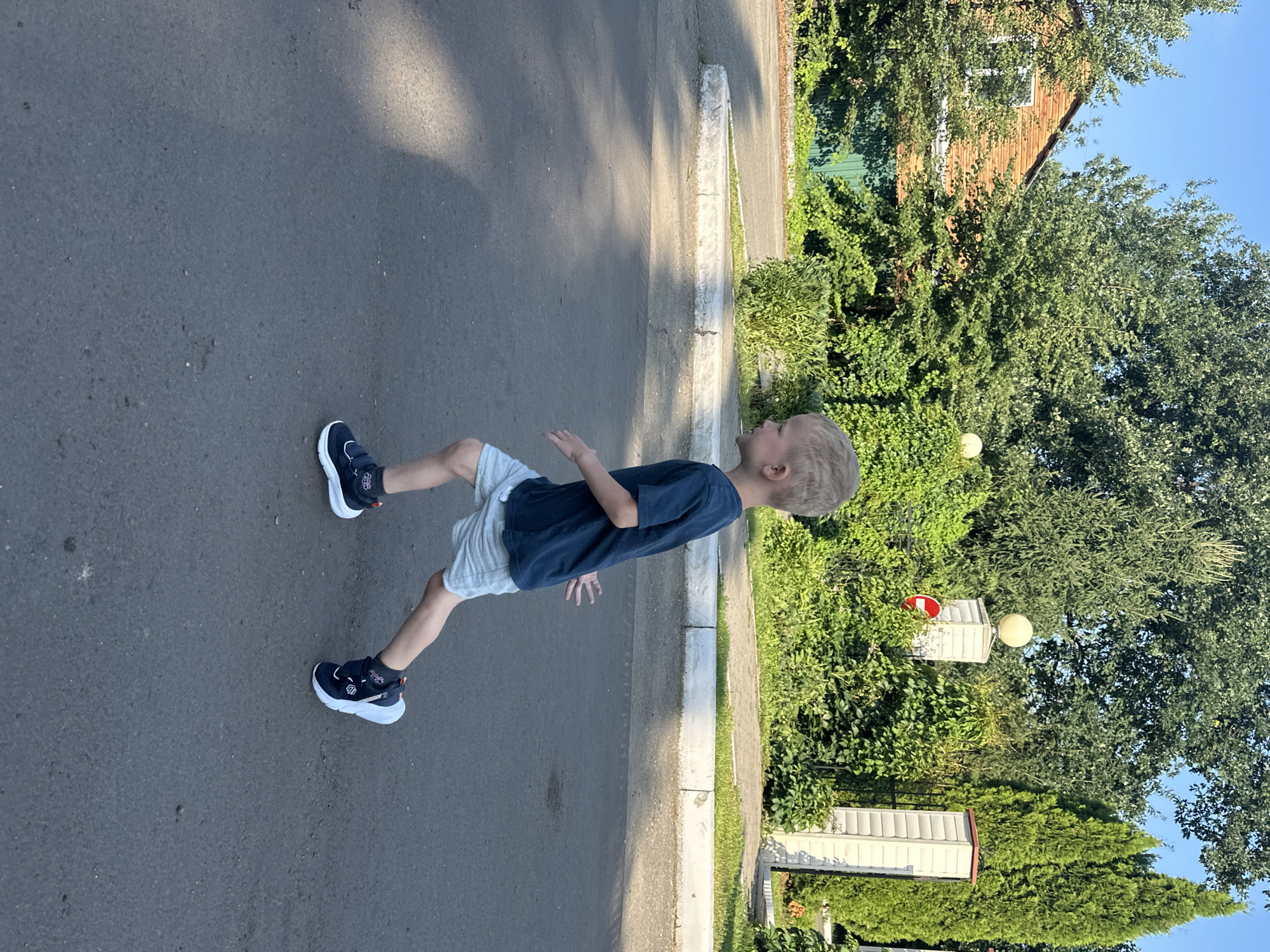 A young boy with blonde hair runs on a paved road in an outdoor residential area. He is wearing a dark blue t-shirt, light blue shorts, and dark blue sneakers with white accents. The boy is positioned in the center of the frame, facing right, with his arms extended slightly. In the background, there is a row of trees and greenery, a wooden house with a green fence, two white pillars with lamps, and a white building with a red and white sign. The ground is asphalt, and a concrete curb runs along the edge of the road. Sunlight casts shadows on the road. The location is in Danilovka, Altai Krai, Russia, likely a suburban or rural setting.