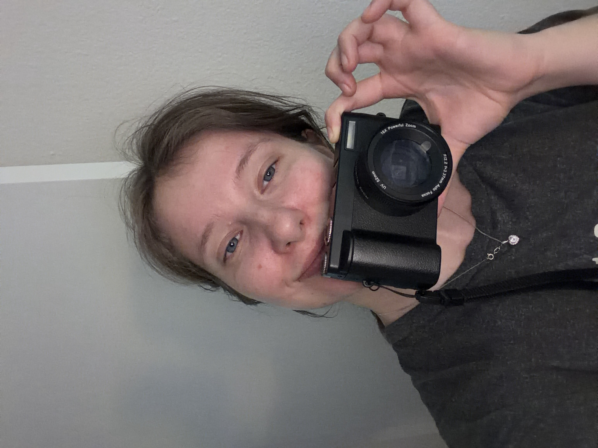 A young woman with light brown hair and blue eyes is holding a black digital camera to take a selfie. She is wearing a dark gray t-shirt and a silver necklace with a heart-shaped pendant. The background is a plain, light gray wall. The image is a medium shot, with the camera lens partially obscuring her mouth and chin. Her left hand is holding the camera, while her right hand is raised and her thumb and index finger are extended, forming a gesture. The lighting is soft and diffused, creating a casual and intimate atmosphere. No specific location in Portland, United States of America, can be determined from the image.