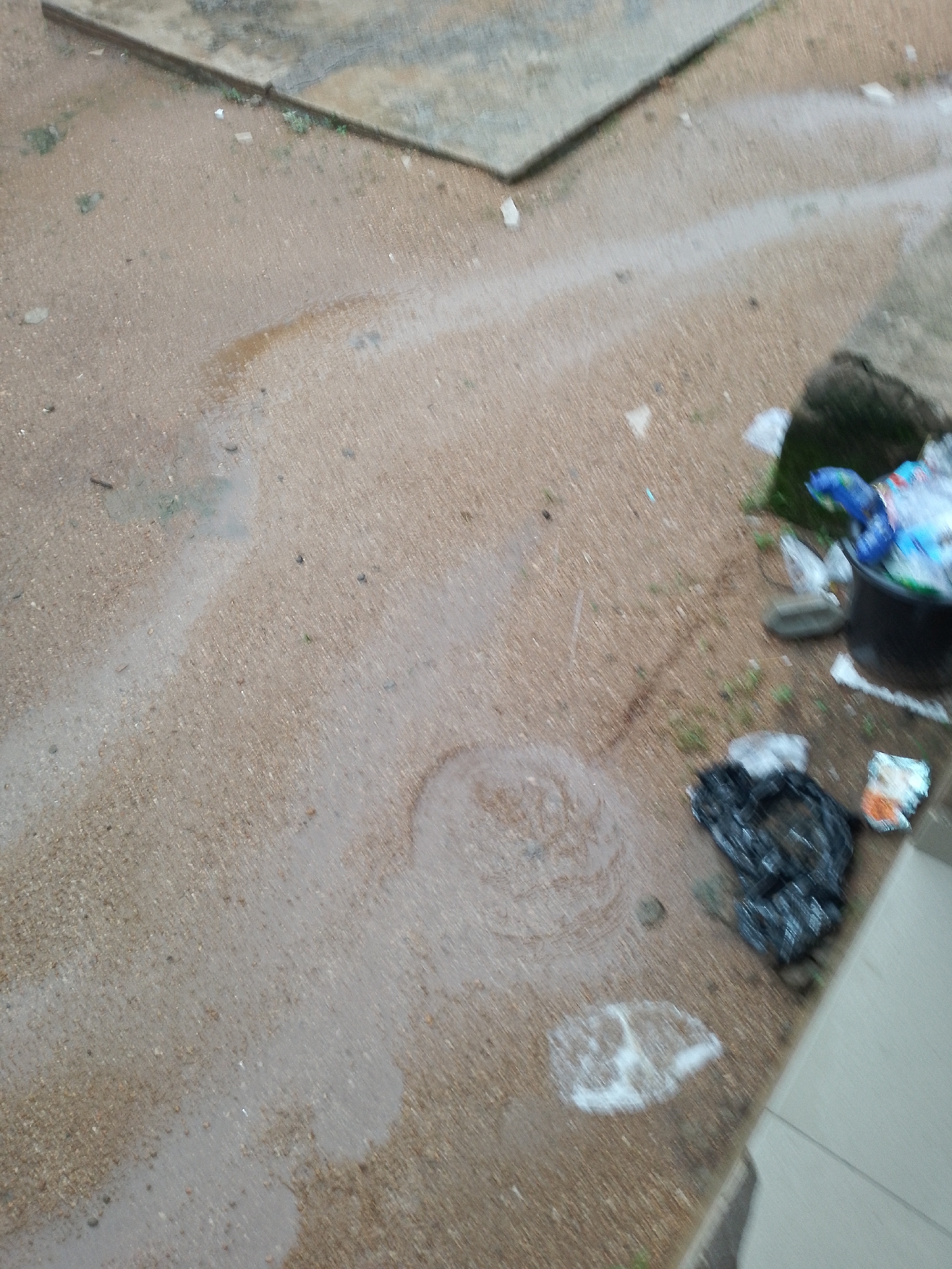 The image depicts a ground-level view of a wet, outdoor environment, likely a walkway or paved area, possibly in a city. There are puddles and streaks of water on the sandy-colored ground, suggesting recent rain. Scattered debris is visible, including plastic bags, wrappers, and what appears to be a partially filled trash bin on the right side of the image. The bin is dark and contains a mix of items, some of which are brightly colored plastic. One black trash bag lies on the ground near the bin. A circular ripple pattern is visible in a puddle on the ground. In the upper left portion of the image, a raised, concrete-like structure is partially visible, with a dark edge. The overall impression is one of a somewhat neglected outdoor space after rainfall.