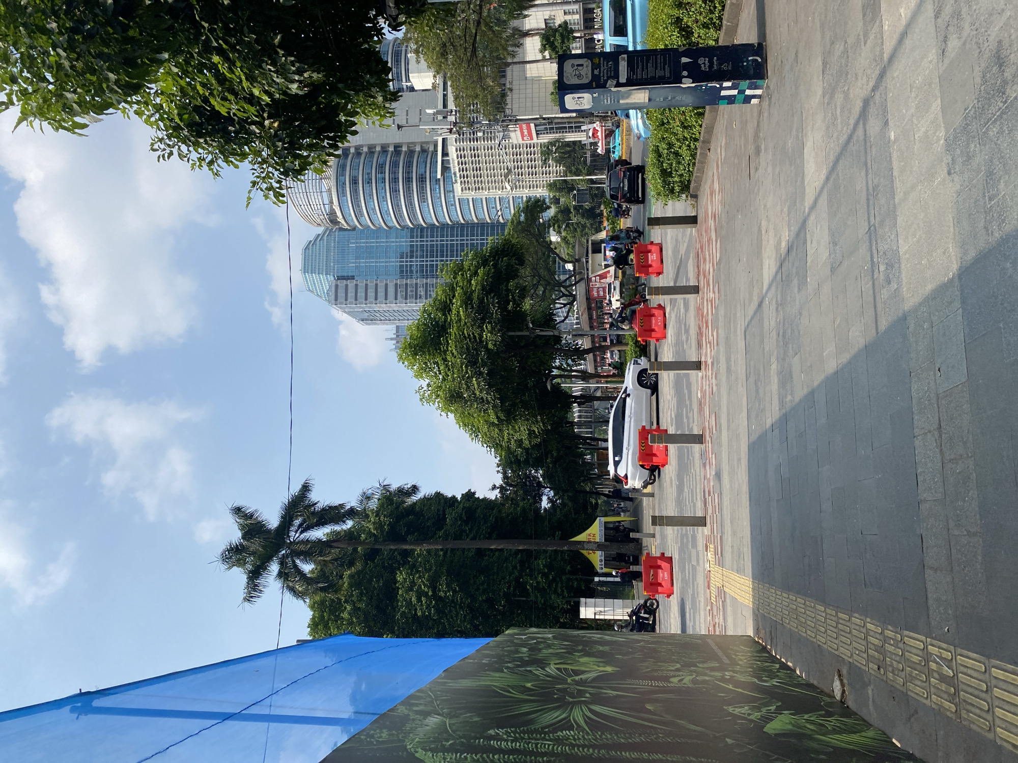 The image shows a street scene in South Jakarta, Indonesia, on a sunny day with a clear sky and scattered clouds. In the foreground, a paved pedestrian walkway with tactile paving for the visually impaired runs along the left side. A large blue construction barrier with a graphic of tropical plants is present on the far left. The walkway leads towards a street with several vehicles, including a white SUV, a black sedan, and a light blue vehicle. Red traffic barriers are placed on the road.
In the midground, lush green trees line the street, with a tall palm tree on the left. Behind the trees, modern skyscrapers dominate the skyline, some with distinctive curved architecture. A yellow tent structure is visible in the distance, with people gathered around it. Various street signs and poles are also present, including one with bicycle lane information. The overall environment suggests a bustling urban area with a mix of greenery and modern infrastructure.