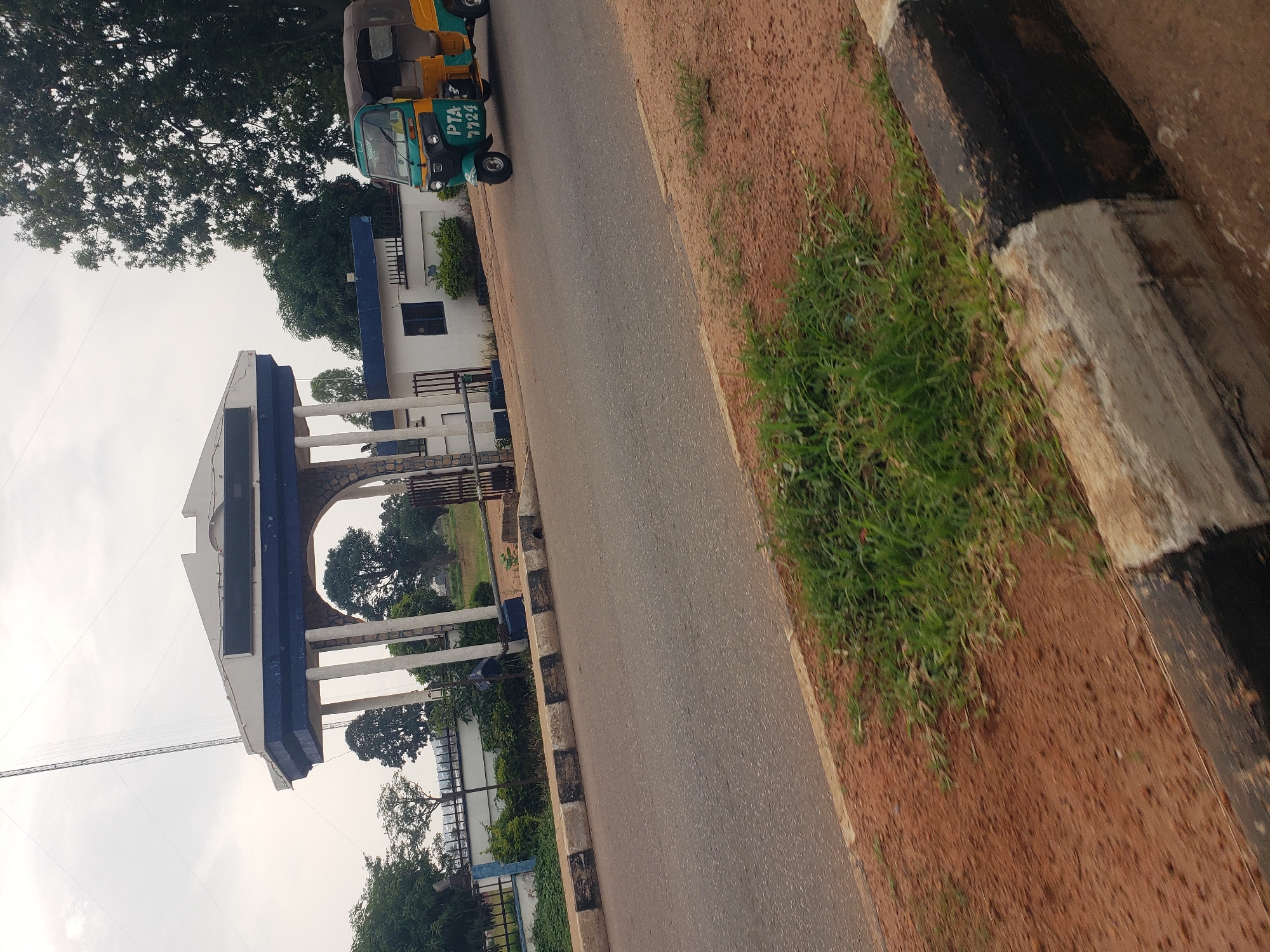 The image captures a scene in Gura Top, Nigeria, featuring a wide asphalt road in the foreground with a red dirt shoulder and some overgrown grass. A concrete curb, painted with alternating black and white stripes, separates the road from the shoulder. In the background, an arched gateway structure stands prominently. This gateway is white with a blue trim and appears to be made of stone and concrete, supported by large white pillars. Beyond the gateway, a white building with dark-framed windows is visible, along with metal fencing. A bright yellow and green auto-rickshaw, with the license plate "PTA 7724", is parked on the road to the right of the gateway. Trees are scattered throughout the background, and a tall antenna structure is visible in the upper left corner. The sky is overcast with a pale, diffused light.