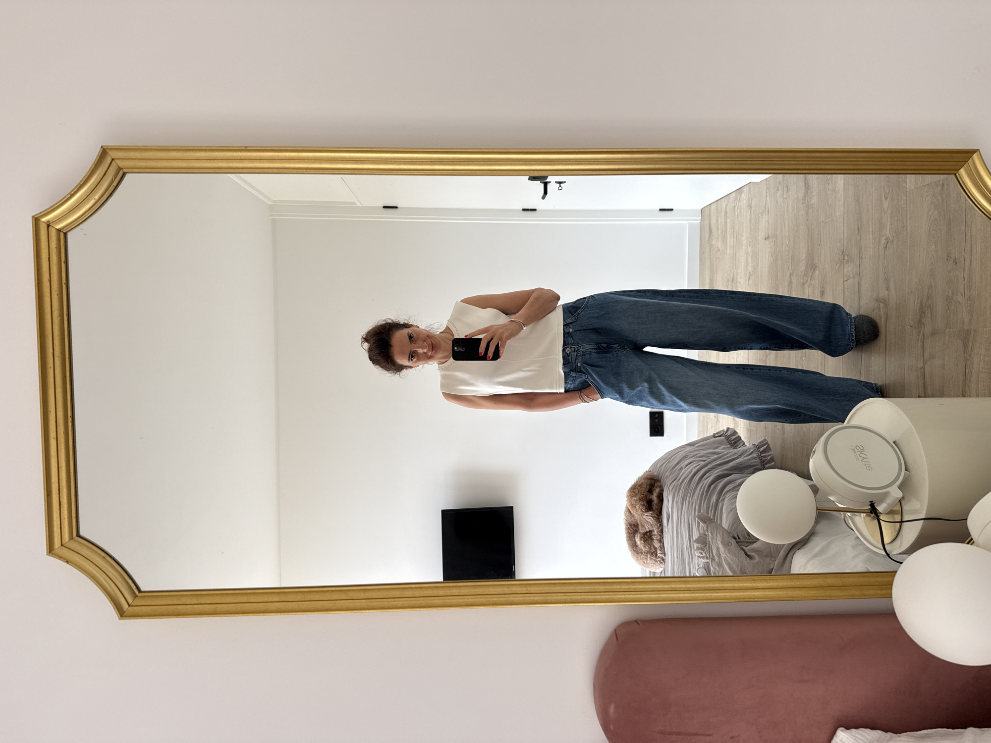 A full-length, ornate gold-framed mirror captures a woman taking a selfie in what appears to be a bedroom or dressing area in Vila Nova de Gaia, Portugal. The mirror, with its decorative, scalloped top corners, is mounted on a plain white wall. The woman stands facing her reflection, holding a black smartphone to take the picture, and smiles faintly. She has dark hair pulled back and is wearing a white sleeveless top and blue wide-leg jeans, with a subtle gold bracelet visible on her right wrist.
The reflection reveals an interior space with light wooden flooring and white walls. In the background, to the right of the woman, a tall, white cylindrical side table holds a modern lamp with two spherical white shades. On the table, a white circular device with "axLite" and "by akko" printed on its top is visible. Next to the table, a rumpled gray textile, possibly bedding, is draped over a bed or seating area, where a small, fluffy brown animal (likely a dog or pet) is nestled. Further back on the floor, a small black rectangular object, possibly an electronic device, is seen. The lower right corner of the mirror reflects a soft pink upholstered headboard or bench. The bright, even lighting suggests it is daytime.