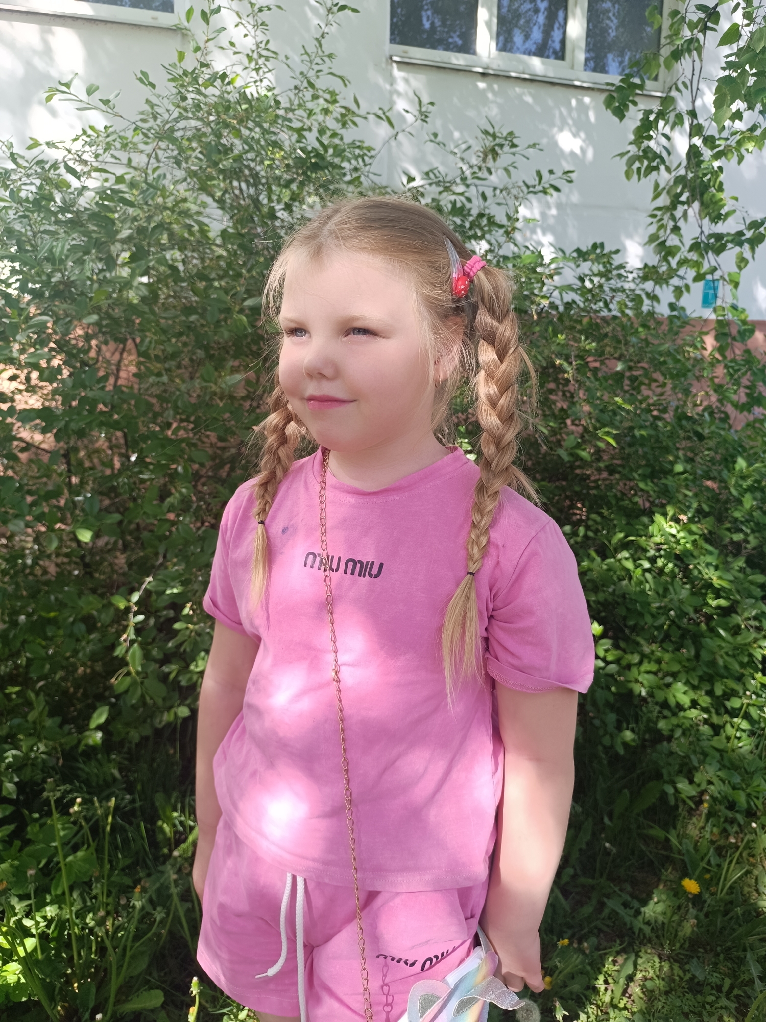 A young girl stands outdoors in front of a lush green bush. She wears a matching pink shirt and shorts with the brand name "MIU MIU" printed on the shirt. Her blonde hair is styled in two braids, held in place by a pink clip on one side. She appears to be holding something with a rainbow effect in her hand. In the background, a white building with visible windows can be seen. The location is Unknown, Unknown.