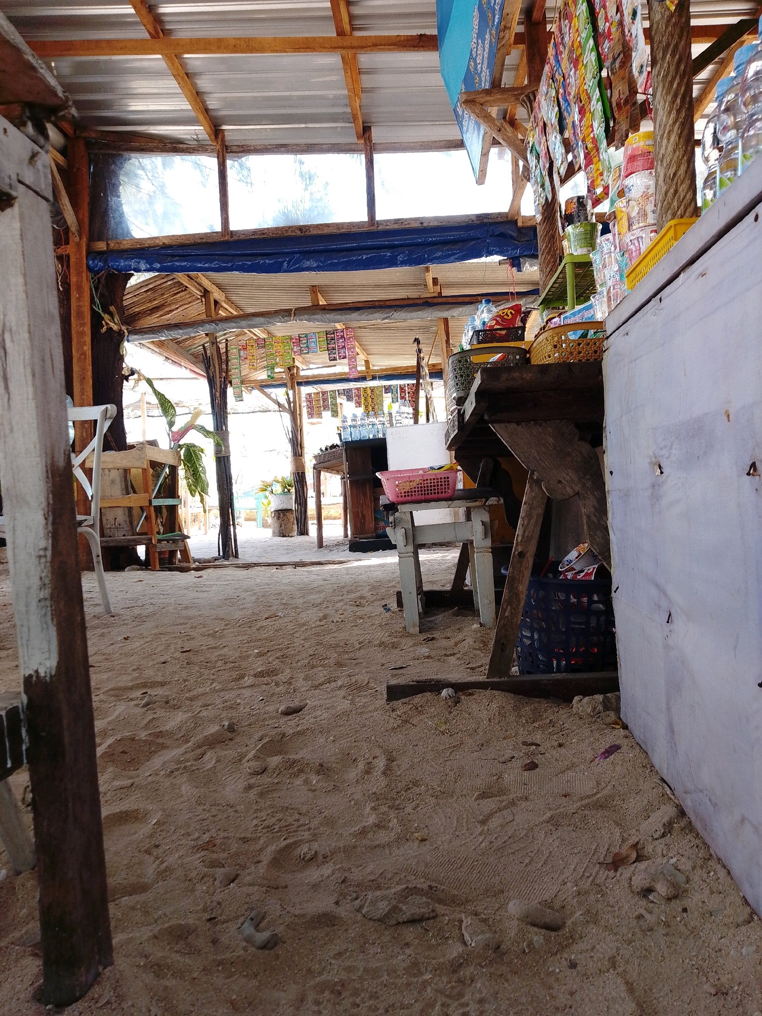 The image depicts an outdoor convenience store situated on a sandy beach in Cikal, Indonesia. The structure is open-air, with a corrugated metal roof supported by wooden beams. The floor is entirely composed of sand, with footprints visible.
Dominating the foreground is a weathered wooden post painted white and dark brown, suggesting it's part of the store's support structure. To the left, a white wooden chair with a distinctive "X" backrest is visible.
The store itself is stocked with various items. A counter area displays rows of colorful snack packets hanging from above. Bottles of water and other beverages are stacked on shelves and within baskets. A pink plastic basket sits on a lower table. Beneath this table, a dark blue woven basket contains more packaged goods.
In the background, more stalls and structures can be seen, with foliage suggesting a tropical environment. Sunlight streams through the gaps in the roofing and walls, illuminating the sandy ground. The overall impression is of a casual, beachside retail establishment.