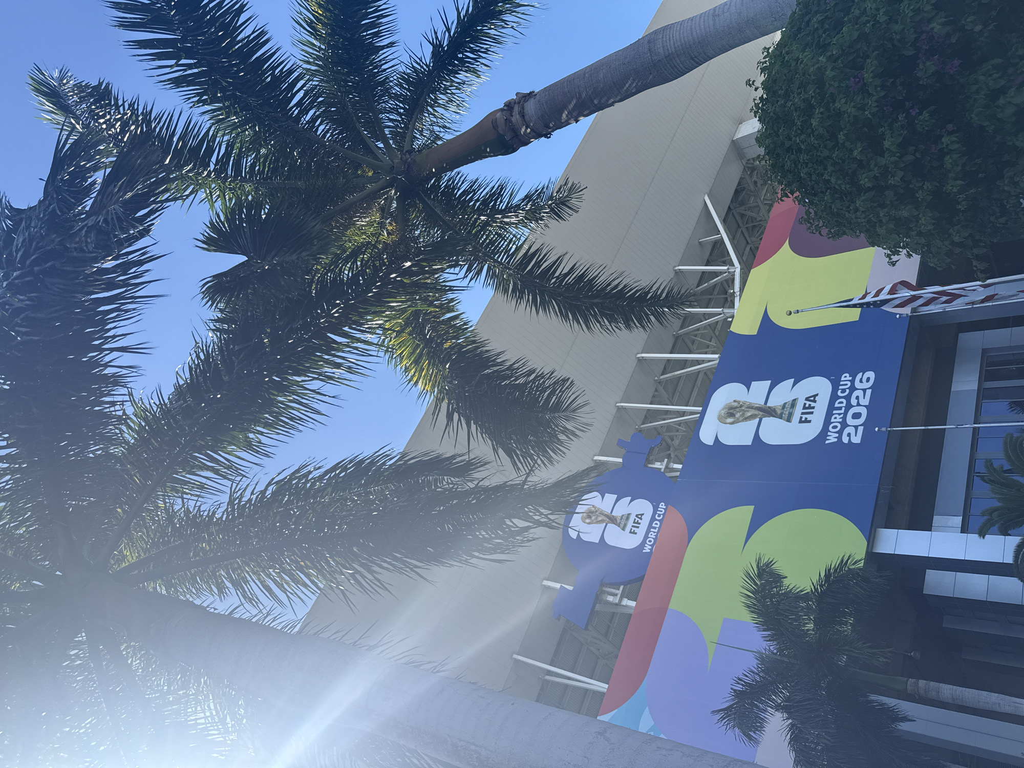 The image shows a vibrant mural for the FIFA World Cup 2026 adorning the exterior of a stadium in Miami Gardens, Florida, United States of America. Two palm trees, one on the left and another on the right, partially obscure the view of the building, their fronds reaching towards a clear blue sky. The mural features bold, colorful graphics, including a large blue section with the FIFA World Cup trophy and the text "FIFA WORLD CUP 2026." The stadium itself has a modern design with a white, angled roof structure. An American flag is visible flying on a flagpole in front of the mural. The scene is bathed in bright sunlight, casting some lens flare into the image.