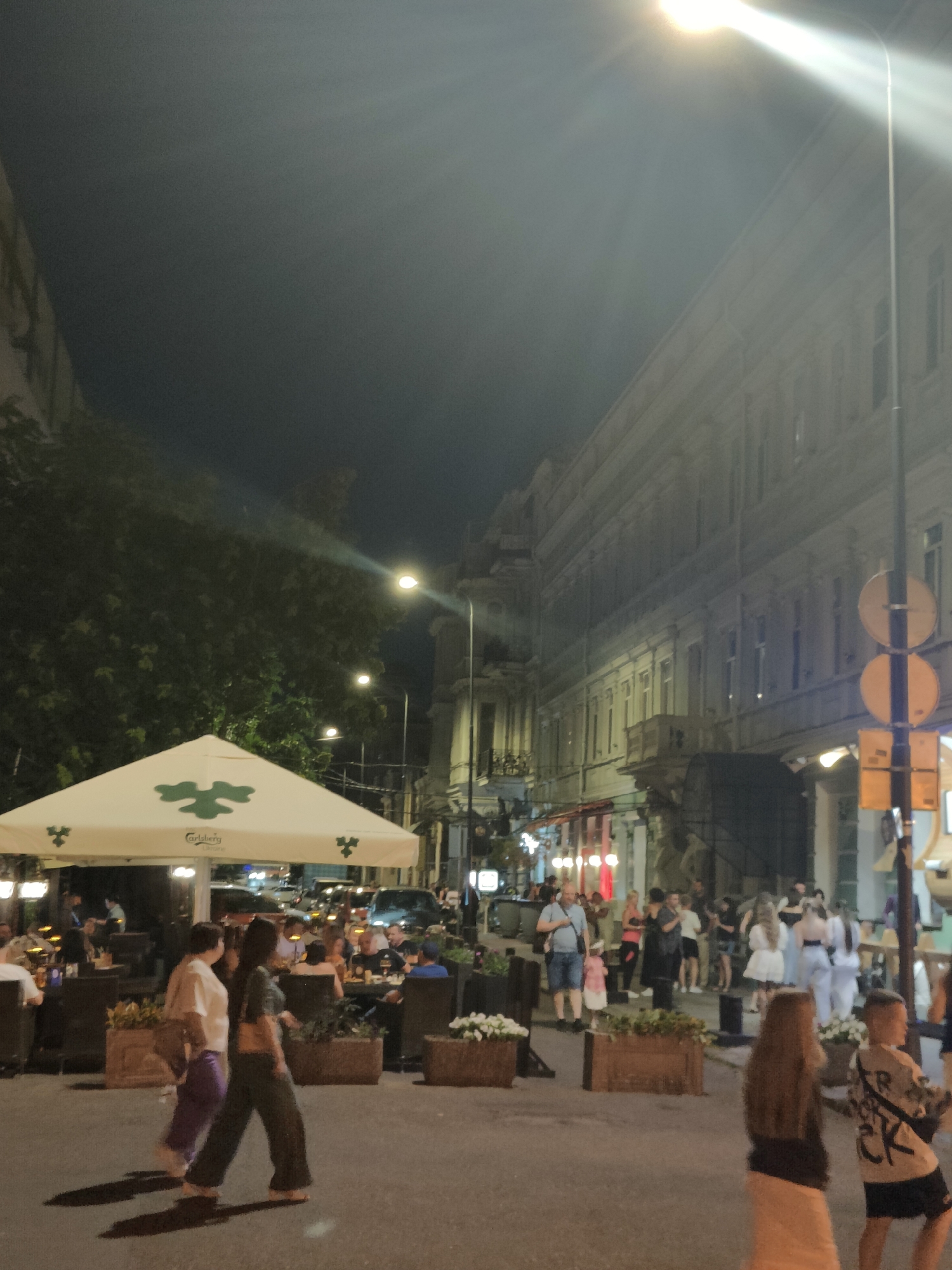 An outdoor street scene at night in Odesa, Ukraine, shows a lively pedestrian area with outdoor dining and groups of people. A large white umbrella with a green logo, possibly indicating a cafe or restaurant, is prominent on the left. Several tables and chairs are set up underneath the umbrella, with patrons seated and dining. In the background, a building with multiple windows lines the street. Streetlights illuminate the scene, casting beams of light and creating a slightly hazy atmosphere. Pedestrians are walking along the street, some in small groups, and others individually. One woman is walking away from the camera, wearing a green top and dark pants. Another woman is walking beside her, wearing a light-colored top and purple pants. A young boy in the foreground is facing away from the camera and appears to be holding something. The overall impression is one of a busy urban night with people enjoying an evening out.