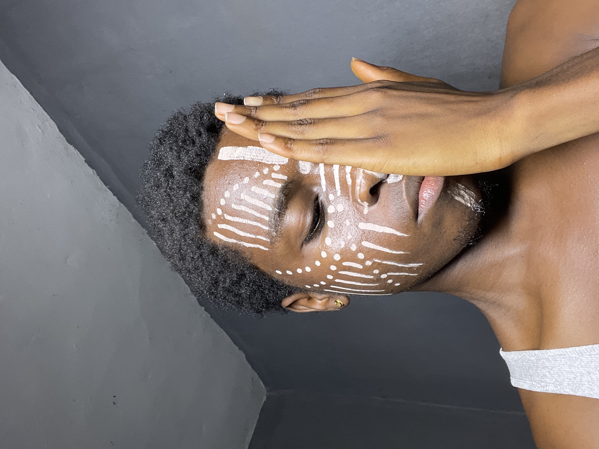 A person with dark skin and curly black hair is presented in a close-up shot. White paint adorns their face in patterns of lines and dots, reminiscent of tribal or cultural markings. Their eyes are closed, and their right hand is held up to their forehead. A gold earring is visible in the person's ear. The background appears to be a dark grey, likely a wall or backdrop. A white strap is visible on their shoulder. The exact location in Nkpor, Nigeria, cannot be determined from the given image.