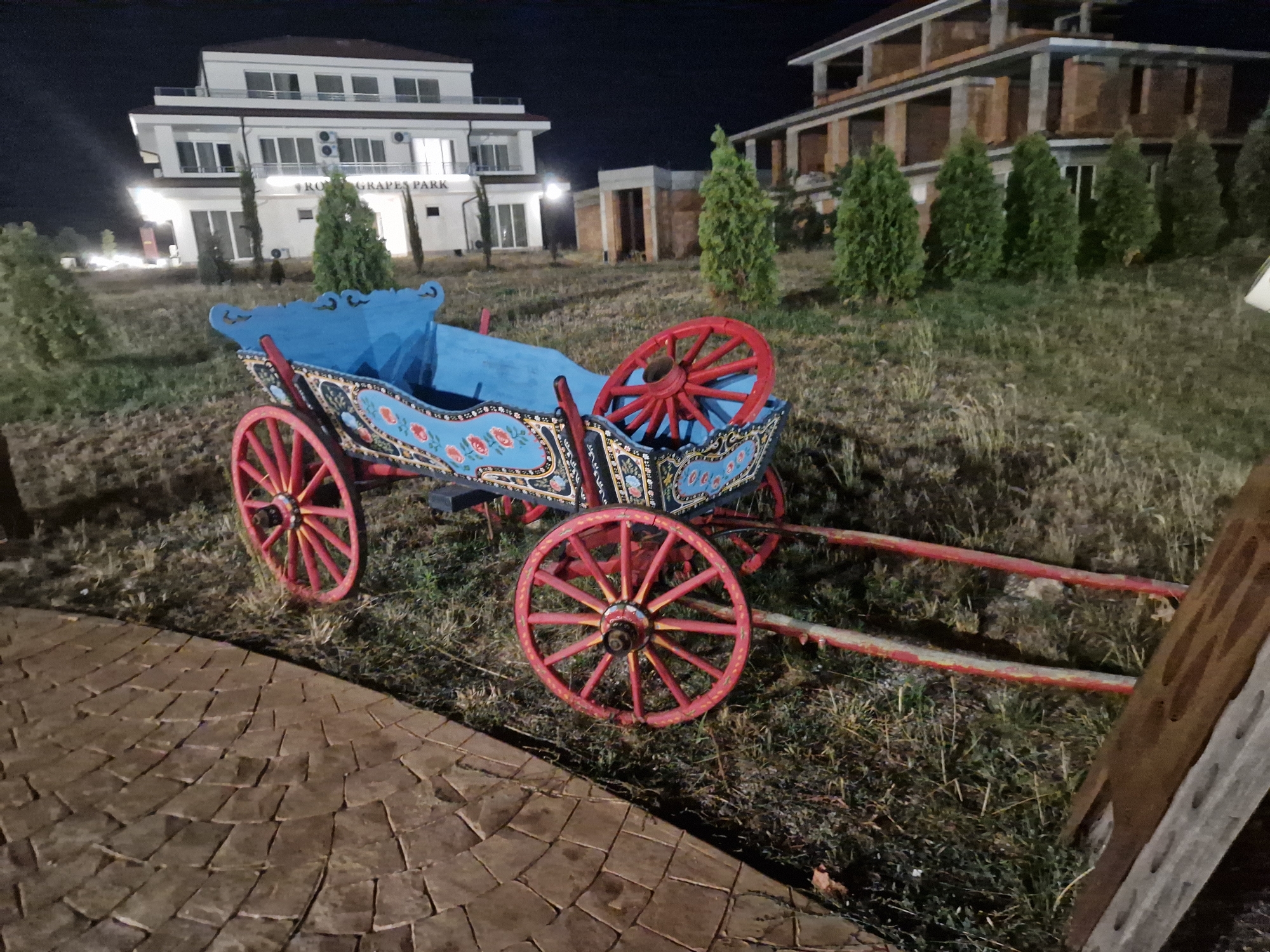 A brightly colored, old-fashioned wooden cart with red wheels and blue trim sits on grassy ground in front of a white building with multiple stories and windows, and a partially constructed building. The cart is ornately decorated with floral patterns in red, blue, and black. The ground in the foreground is paved with irregular brown stones arranged in a circular pattern. Several green evergreen trees are planted in a row behind the cart, and a few more are visible in the background. The sky is dark, indicating it is nighttime. The sign on the white building reads "RO GRAPES PARK". The scene is located in the city of Tankovo, Bulgaria.