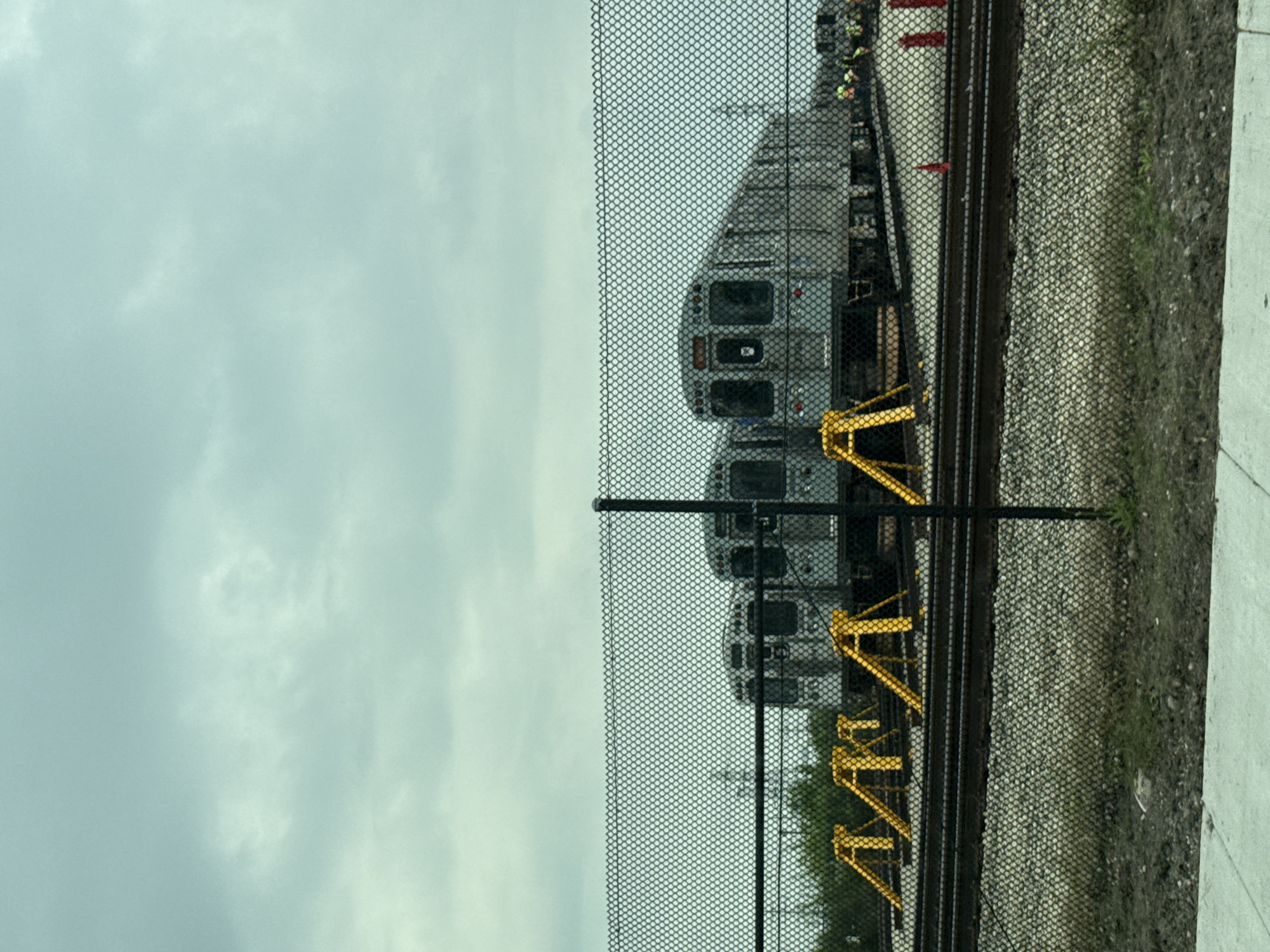 In Skokie, Illinois, United States of America, a chain-link fence encloses a rail yard where several silver commuter trains are parked on tracks. The trains are positioned nose-to-tail, with the leading train's front visible and the others receding into the distance. Yellow wheel chocks are placed on the tracks in front of the trains. The ground is covered with gravel and some patches of dirt and grass. In the background, the sky is overcast with gray clouds.