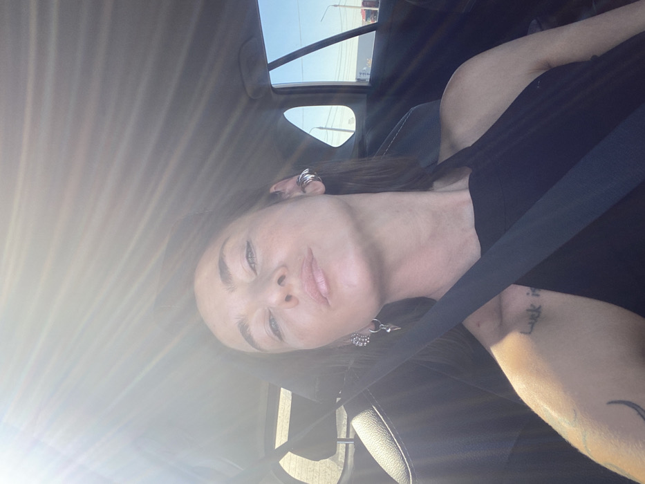 The image displays a female individual seated within a vehicle, secured by a black seatbelt, with the setting identified as the city of Sheragul, Russia. She has fair skin, light-colored eyes, and brown hair, looking directly at the camera with a neutral expression and slightly parted lips. Her attire consists of a black sleeveless top. She wears multiple silver-toned earrings: several hoops/cuffs on her left ear and two distinct pieces on her right ear, one appearing spherical and the other a dangling element with a pointed, conical shape. A black script tattoo, partially legible as "luck is," is visible on her left upper arm, accompanied by a crescent moon-like design. Bright sunlight enters the vehicle from the upper left, causing lens flare and strong illumination. Through the vehicle's side and rear windows, a clear blue sky, distant structures, and power lines are visible, indicating an outdoor, possibly urban or semi-urban, environment during daylight hours.