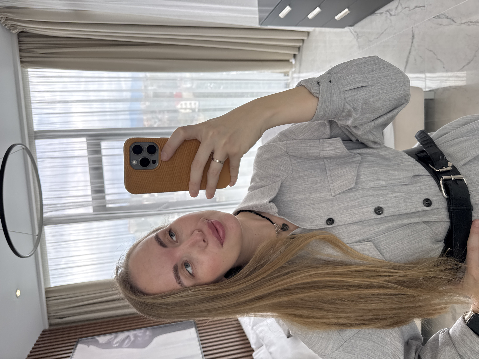 A young woman with blonde hair is taking a selfie indoors. She is wearing a light grey button-down shirt with a black belt. She is holding a brown smartphone with her left hand. The background shows a window with blinds and a partial view of a cityscape, suggesting an urban location. Based on the window view and interior design elements, the location is suggested to be in the city of Sochi, Russia. Visible in the room are modern furnishings, including a white bed with wood headboard and a gray dresser on the wall.